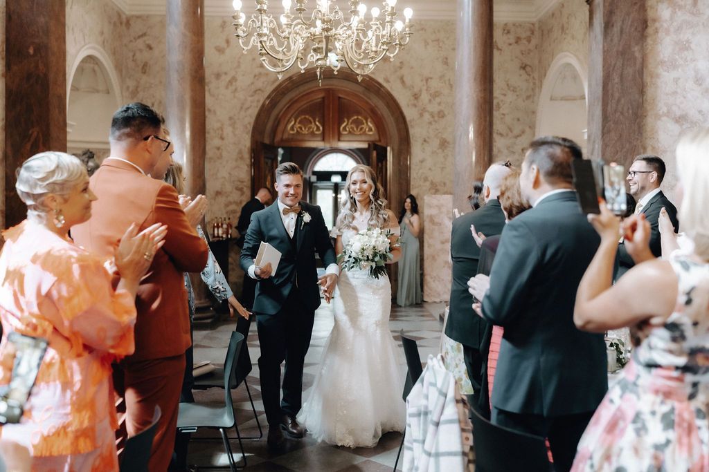 Bride and groom recessional through cheering guests in grand chandelier-lit wedding hall.