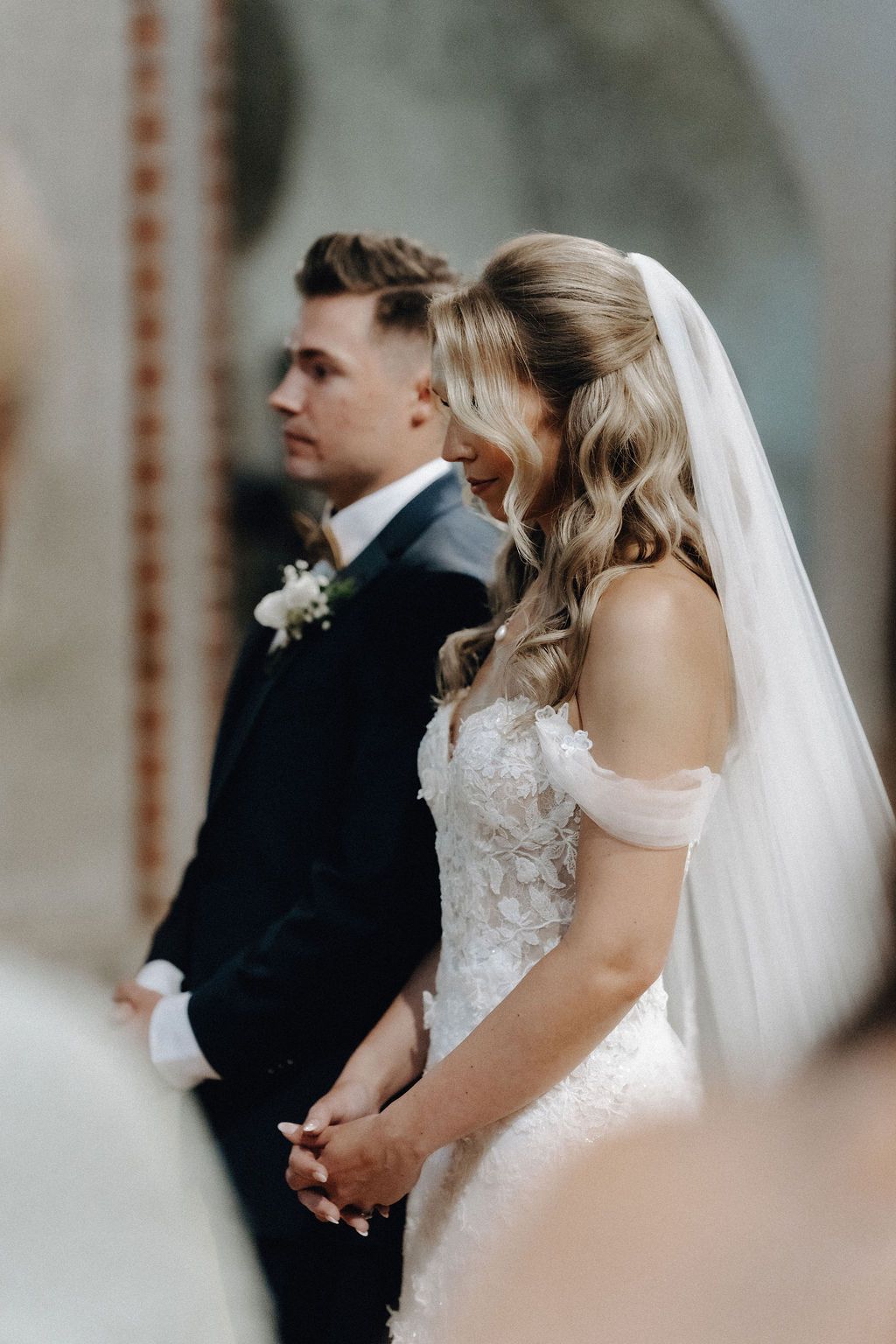 Bride in off-shoulder lace gown and groom stand together praying during church ceremony.