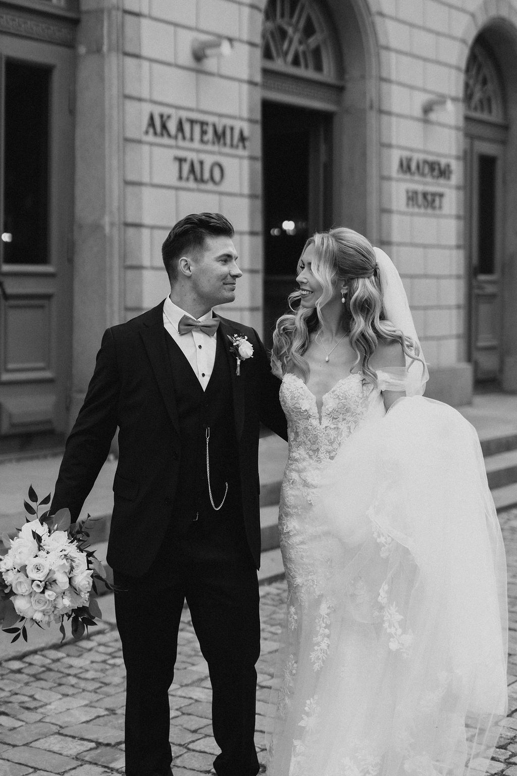 Black and white photo of smiling bride and groom standing outside Akatemia-Talo building on wedding day.