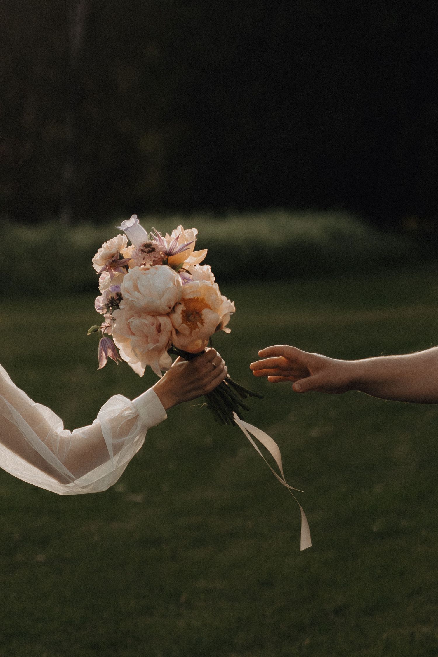 Two hands passing a romantic bouquet of peonies and anemones with ribbon streamers outdoors.