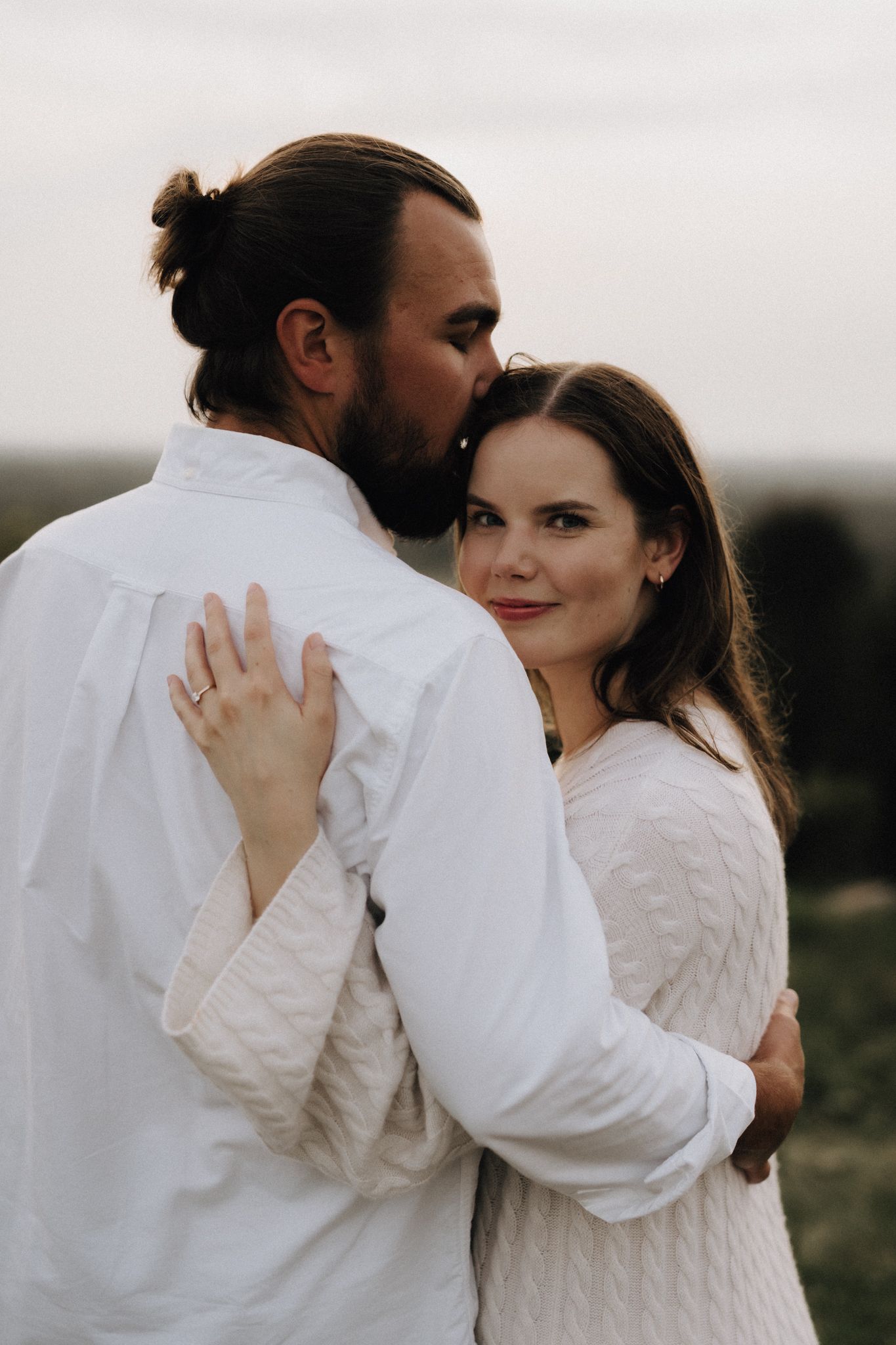 A bearded man with a bun kisses a smiling woman on the cheek as she looks toward the camera outdoors.