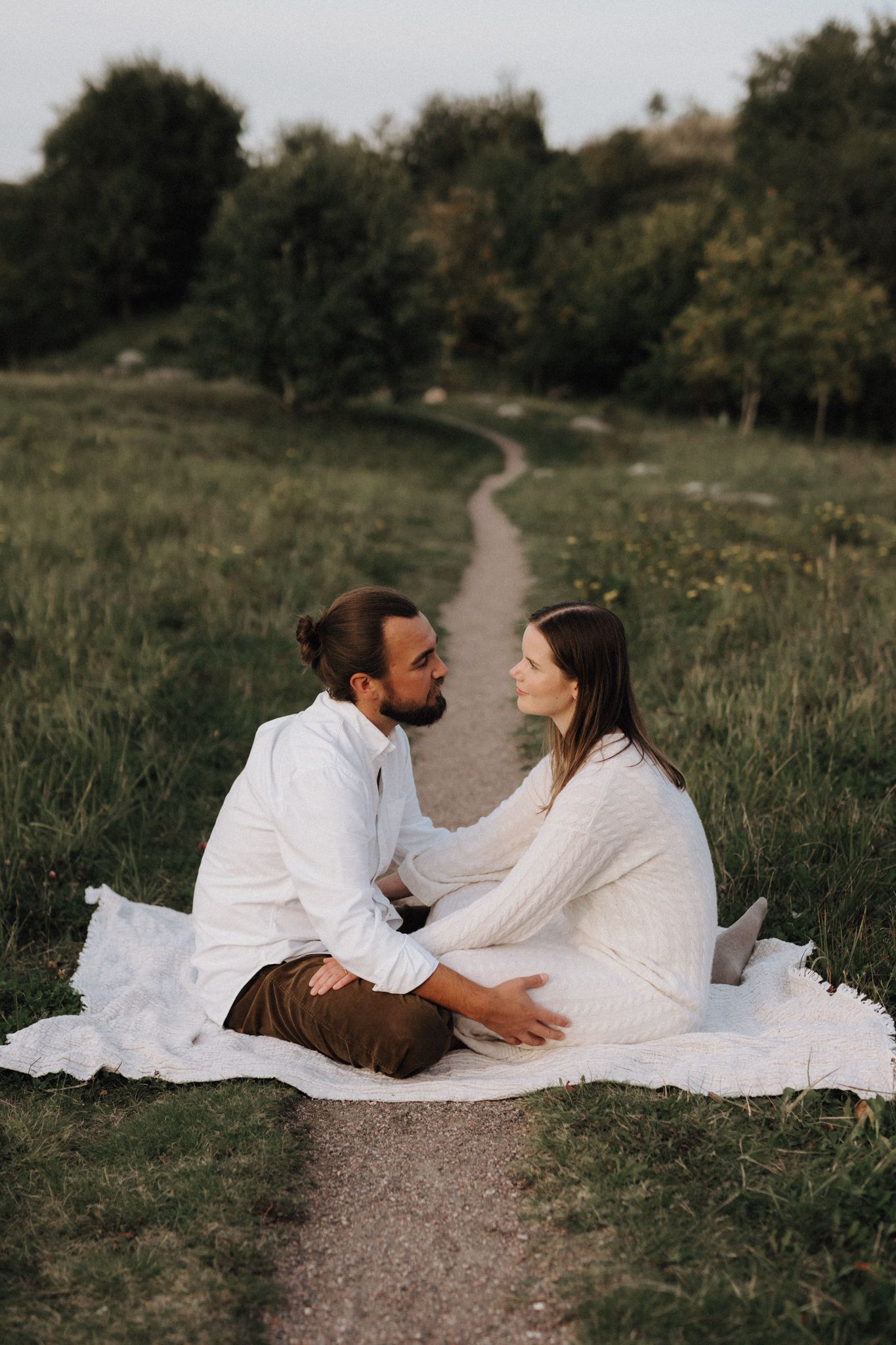 A couple sits facing each other on a blanket on a scenic grassy hillside path.