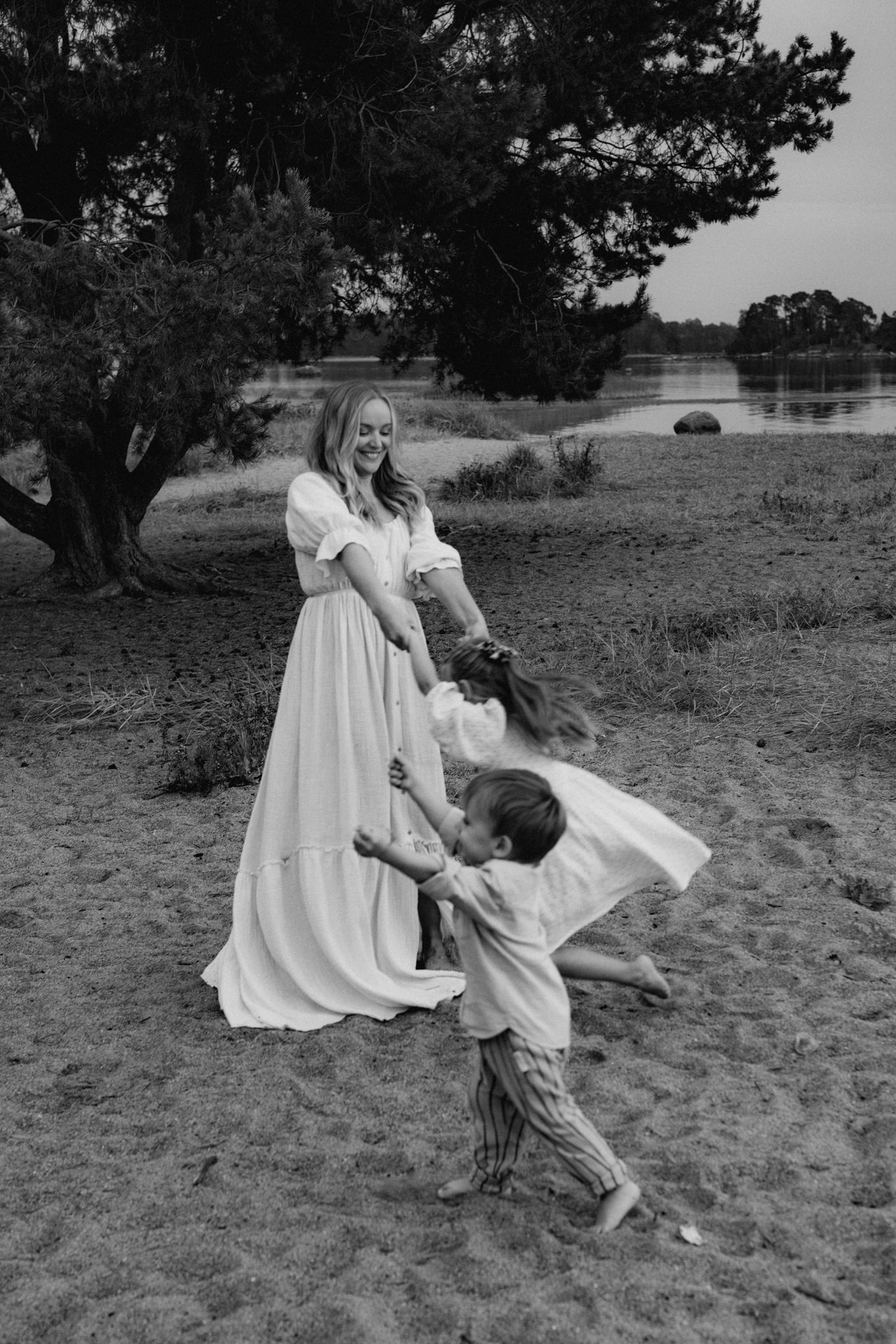 A mother in a white dress plays and spins with her two children on a sandy beach in black and white.