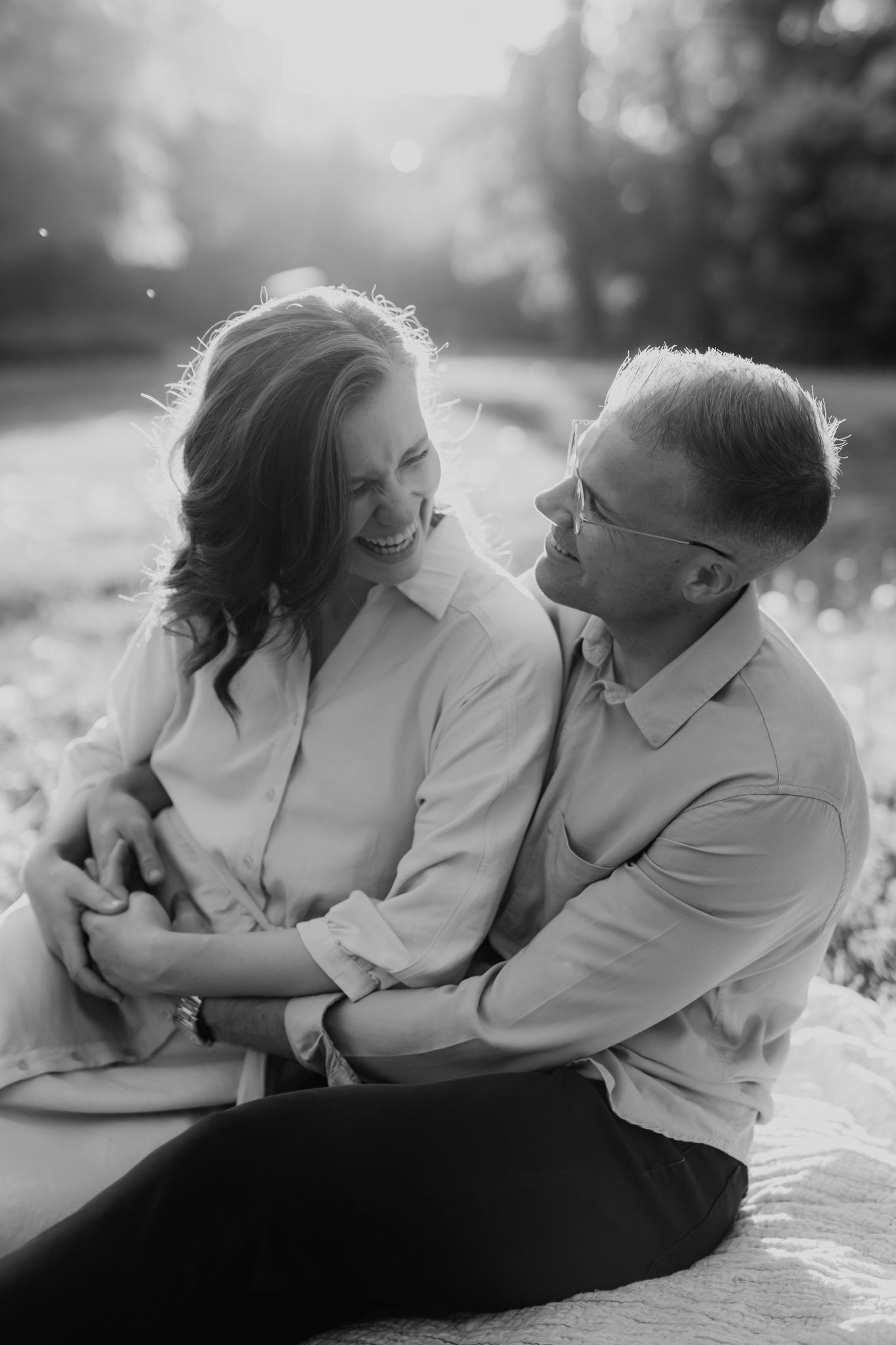 Black and white photo of a couple sitting closely together laughing on a blanket outdoors.