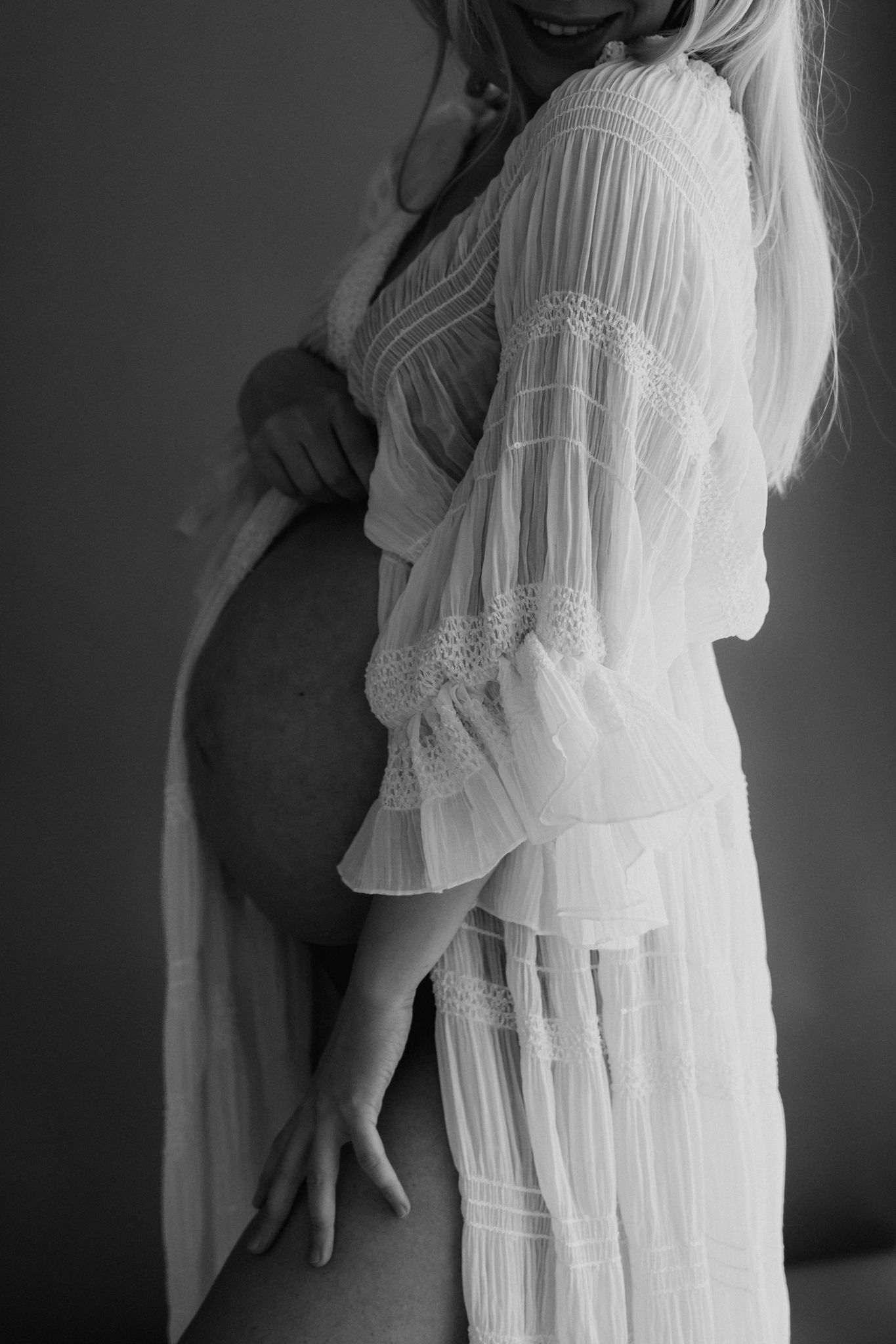 Pregnant blonde woman in flowing white lace dress holding her bare baby bump, elegant black and white maternity photo.