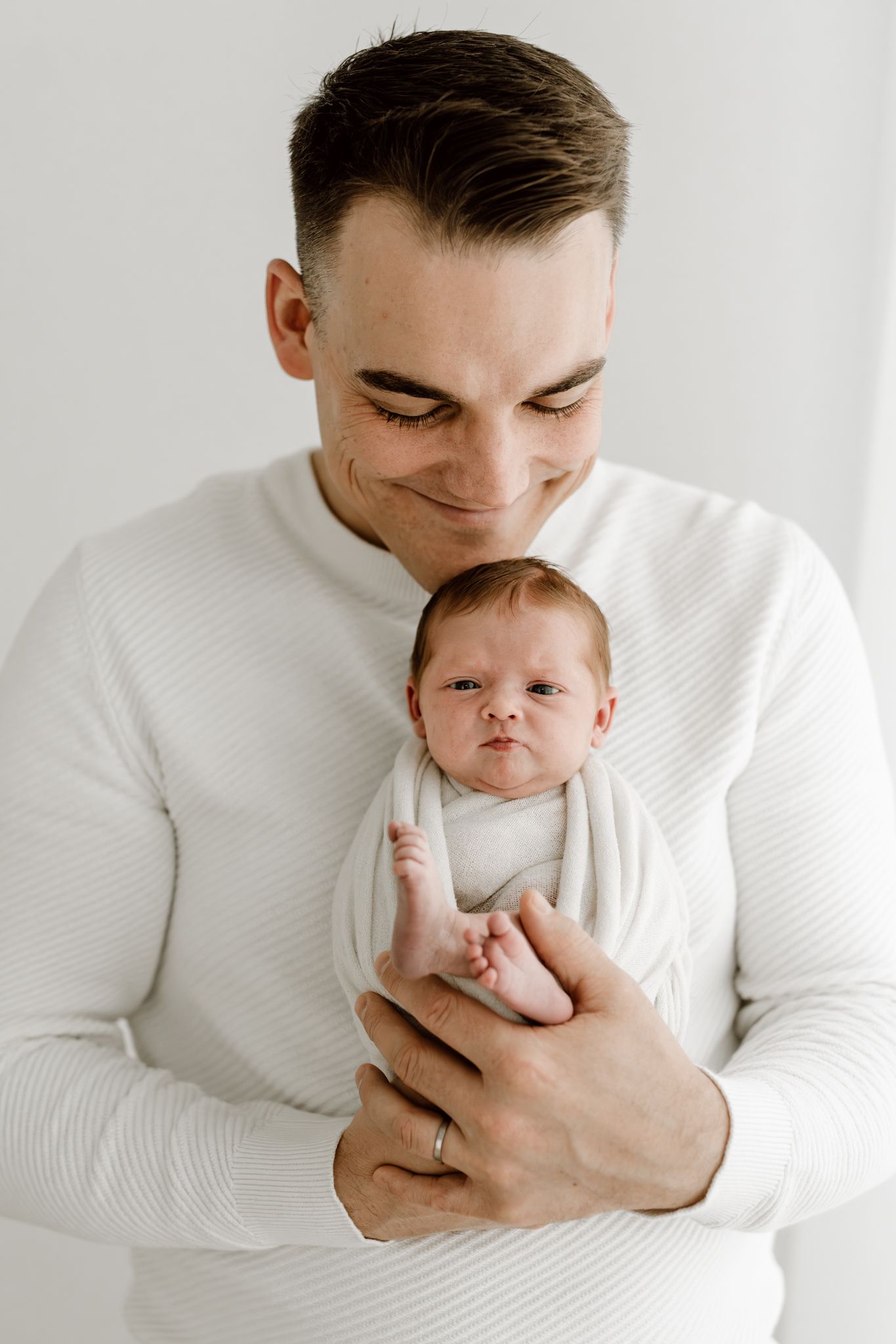 Smiling father in white ribbed sweater holds alert newborn baby wrapped in white swaddle during photo session.