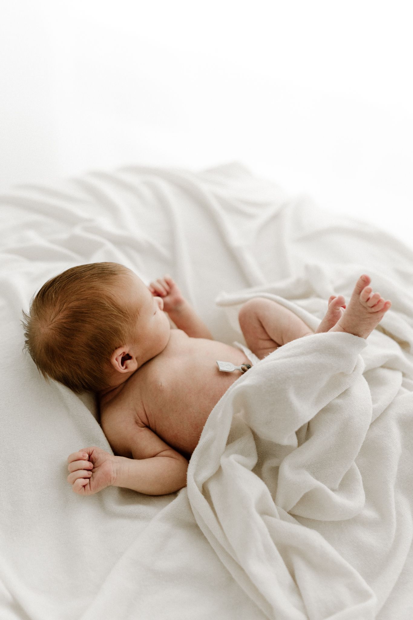 Naked newborn baby lies on white blanket with tiny feet raised during a professional newborn photography session.