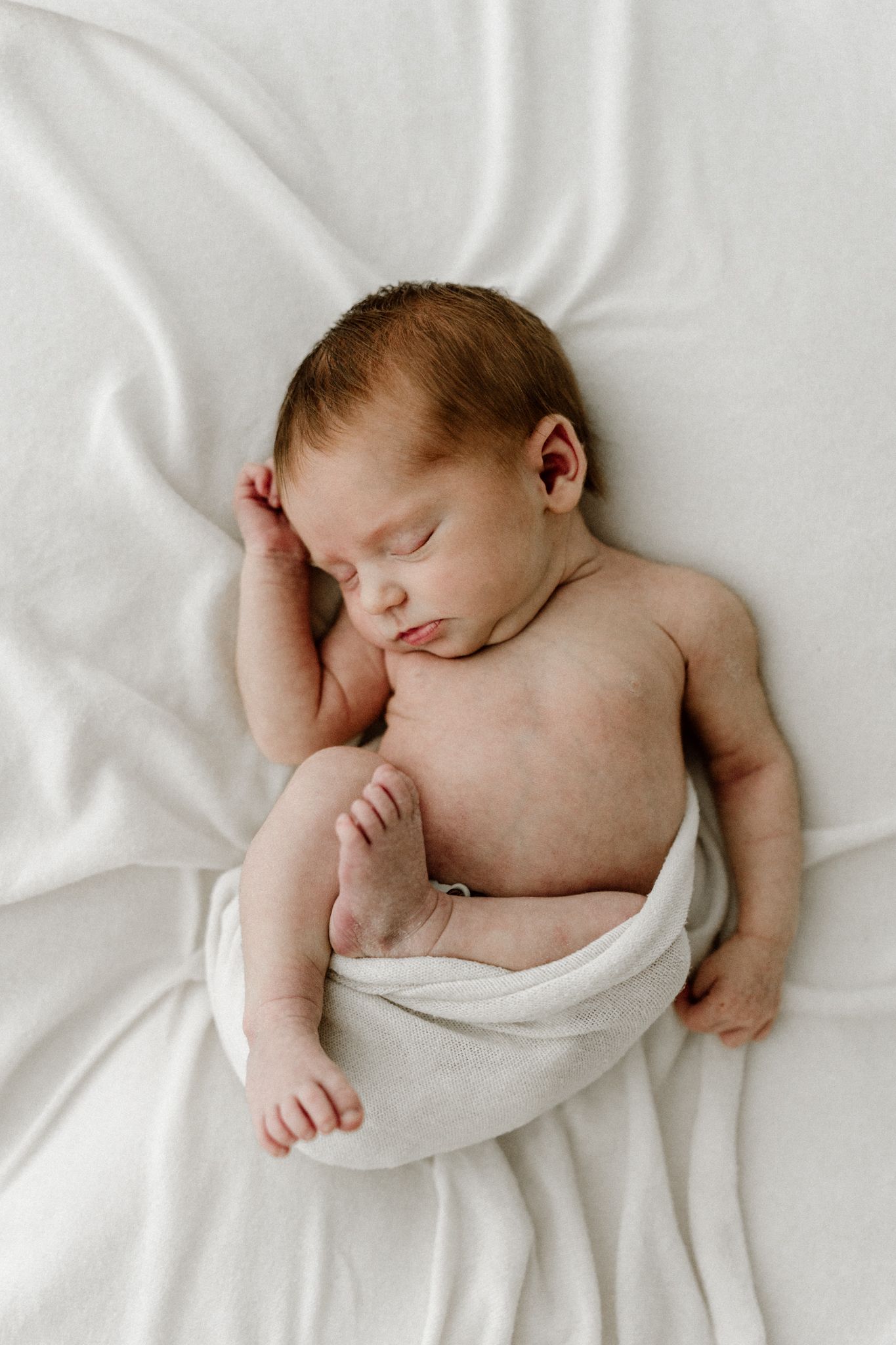 Newborn baby sleeping peacefully on white blanket wrapped in soft white swaddle.