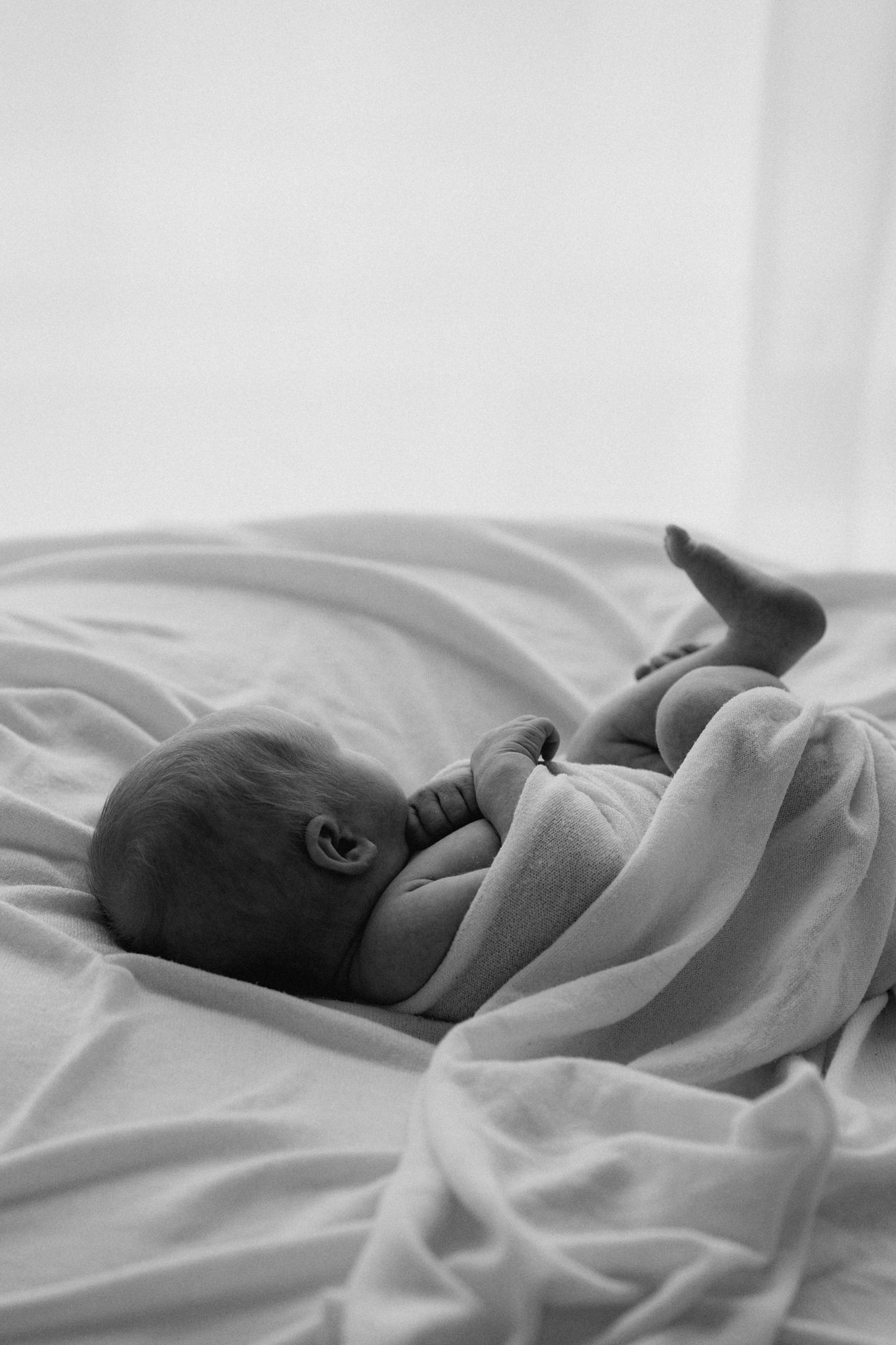 Black and white photo of naked newborn baby lying on white blanket with leg raised.
