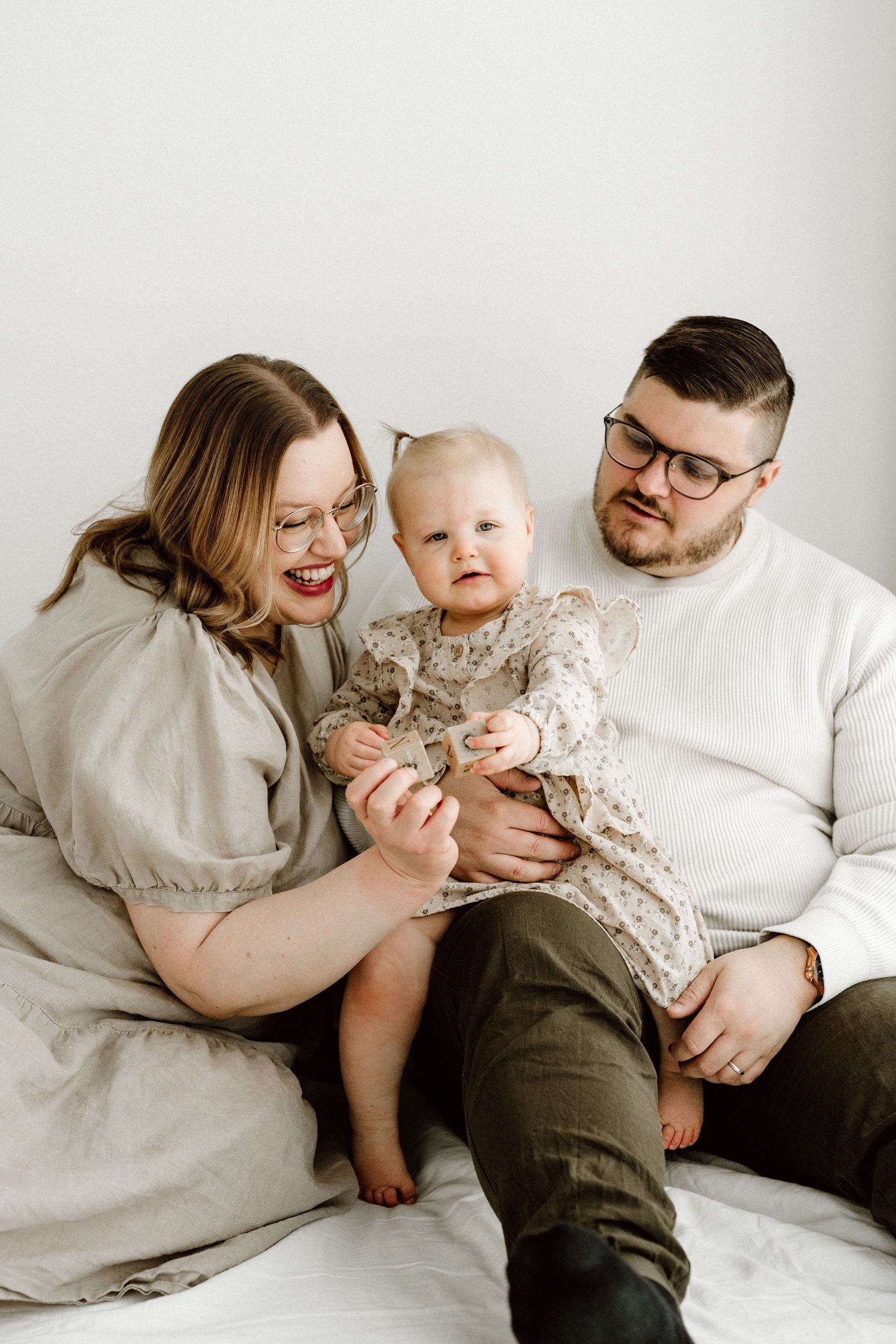A happy couple wearing glasses sits on a white bed holding their baby girl in a floral outfit.