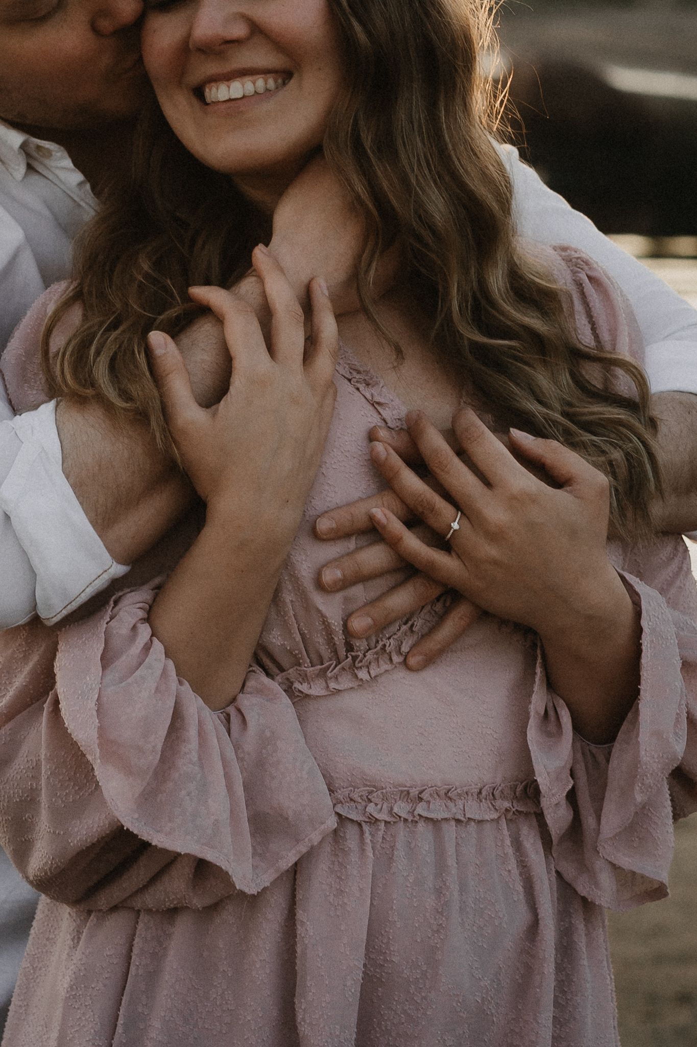 Close-up of couple embracing, woman in pink ruffled dress smiling with engagement ring visible.