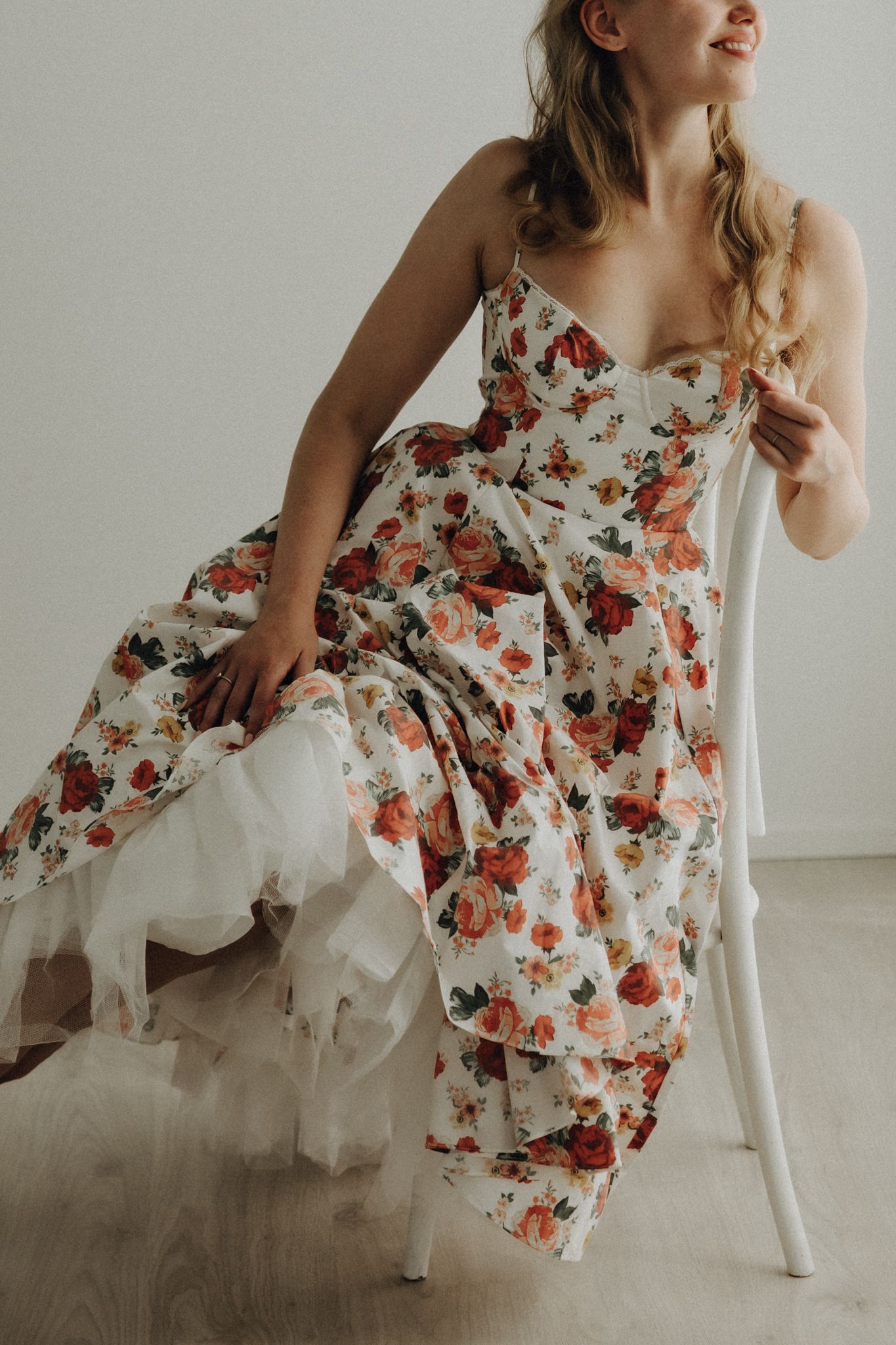 Smiling blonde woman in strapless floral rose print gown sitting gracefully on white chair.