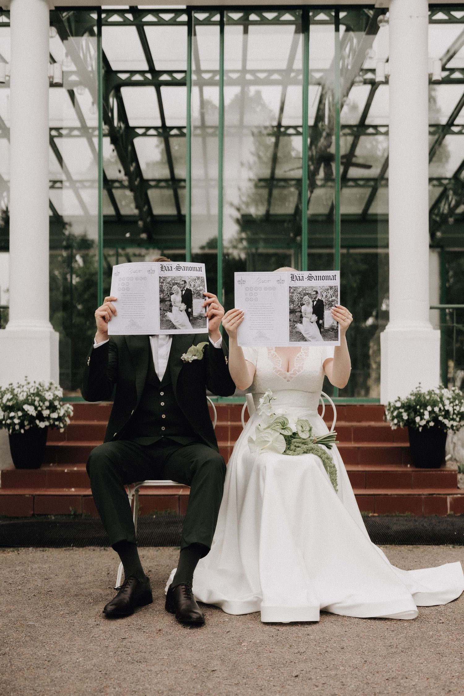 A bride and groom sit holding wedding newspapers covering their faces outside a glass greenhouse venue.