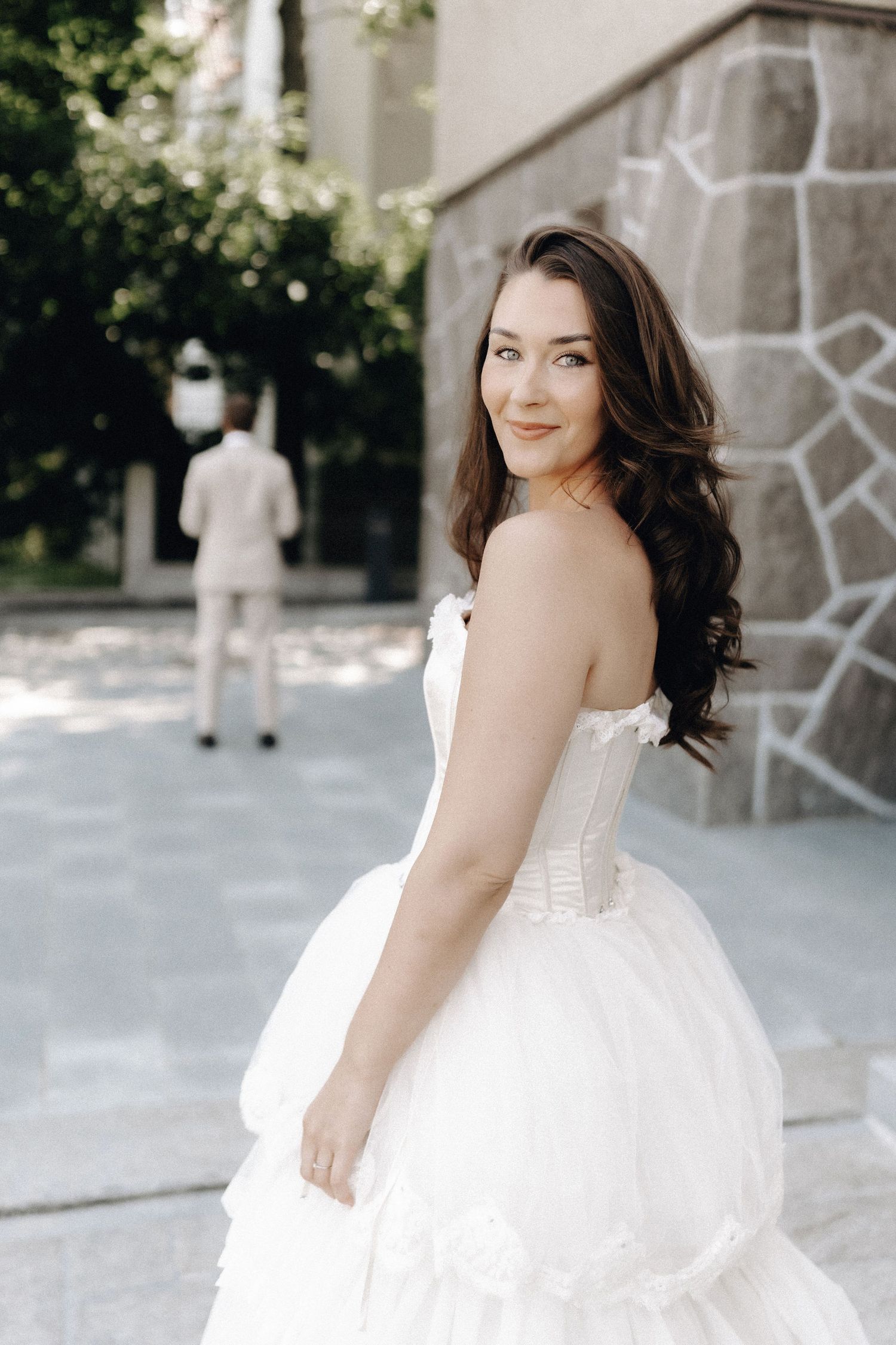 Smiling brunette bride in strapless white ballgown with layered skirt looking back over her shoulder outdoors.