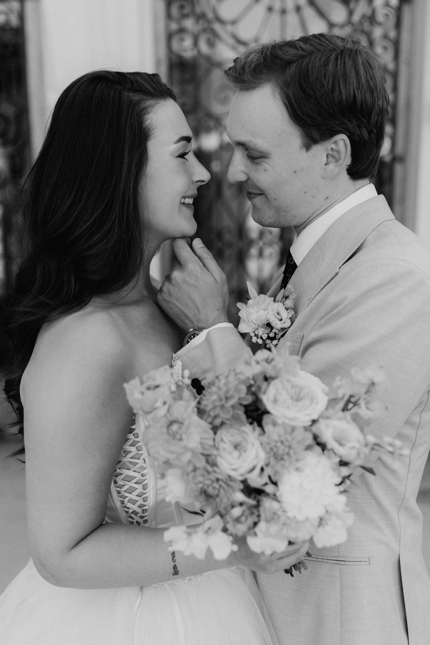 Black and white close-up of smiling bride and groom facing each other holding a floral bouquet.