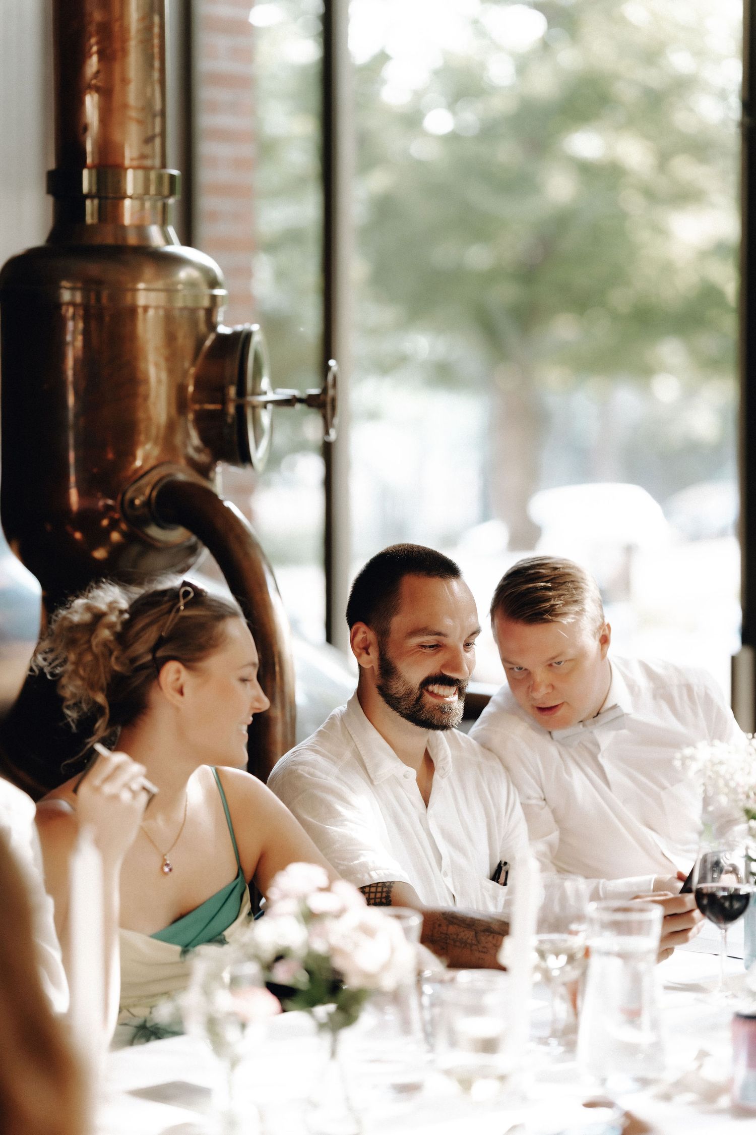 Three wedding guests laugh together at a reception table near a copper distillery vessel.