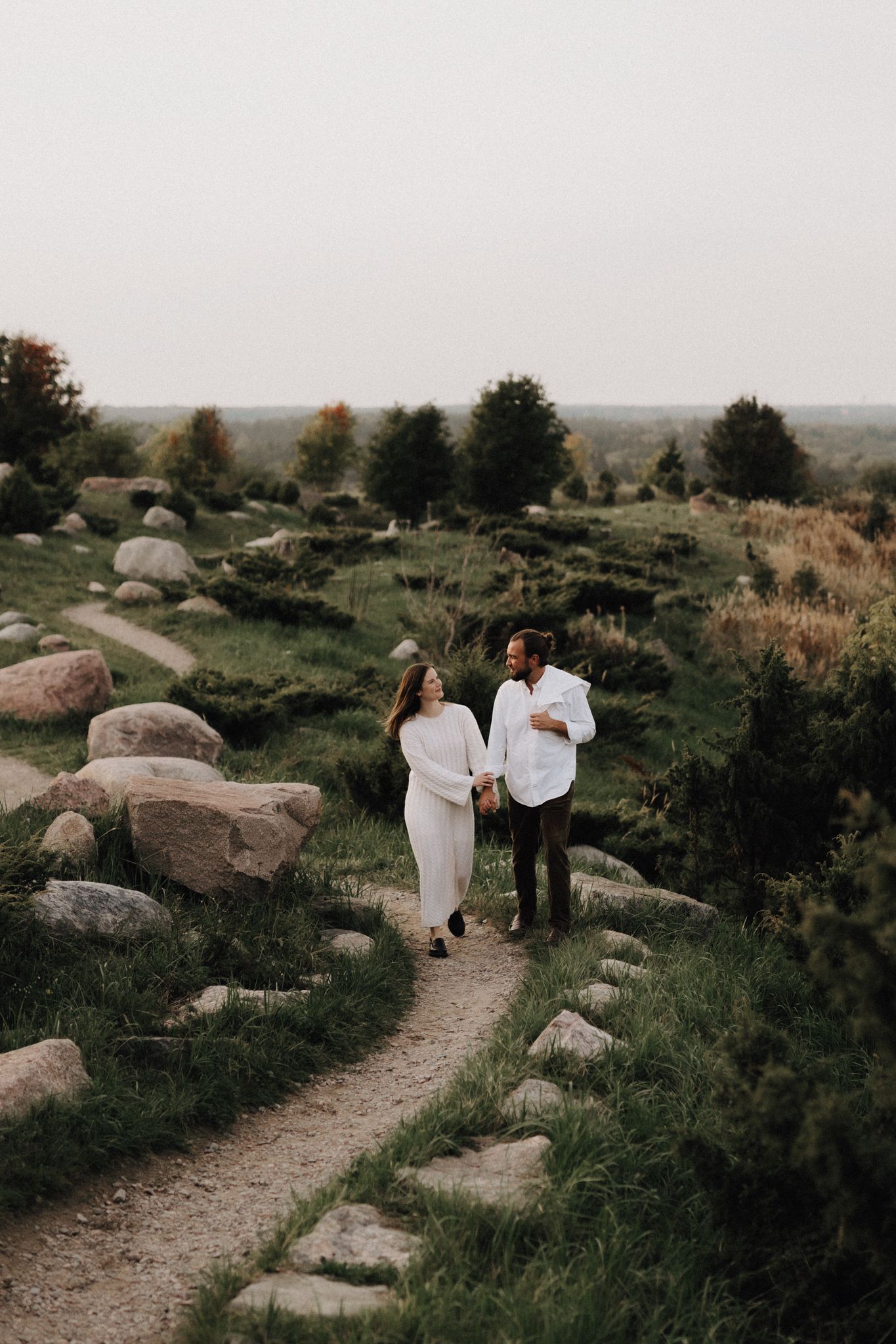 A couple in white outfits holds hands while walking along a scenic rocky garden path surrounded by greenery.