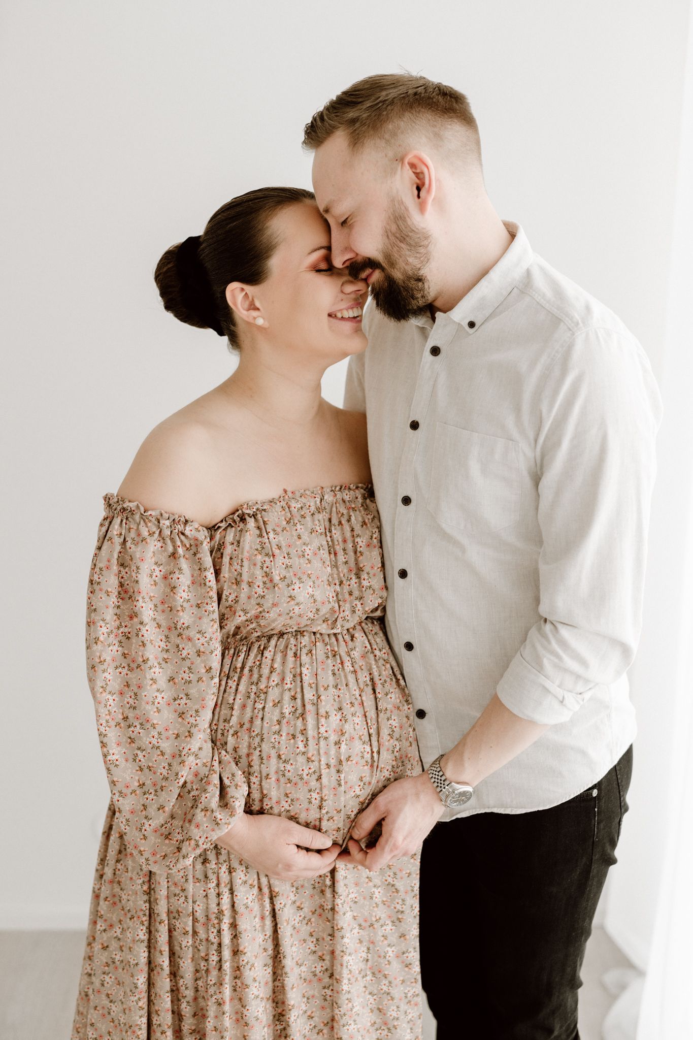 Smiling pregnant couple touching foreheads, woman in floral off-shoulder dress, man in white shirt.