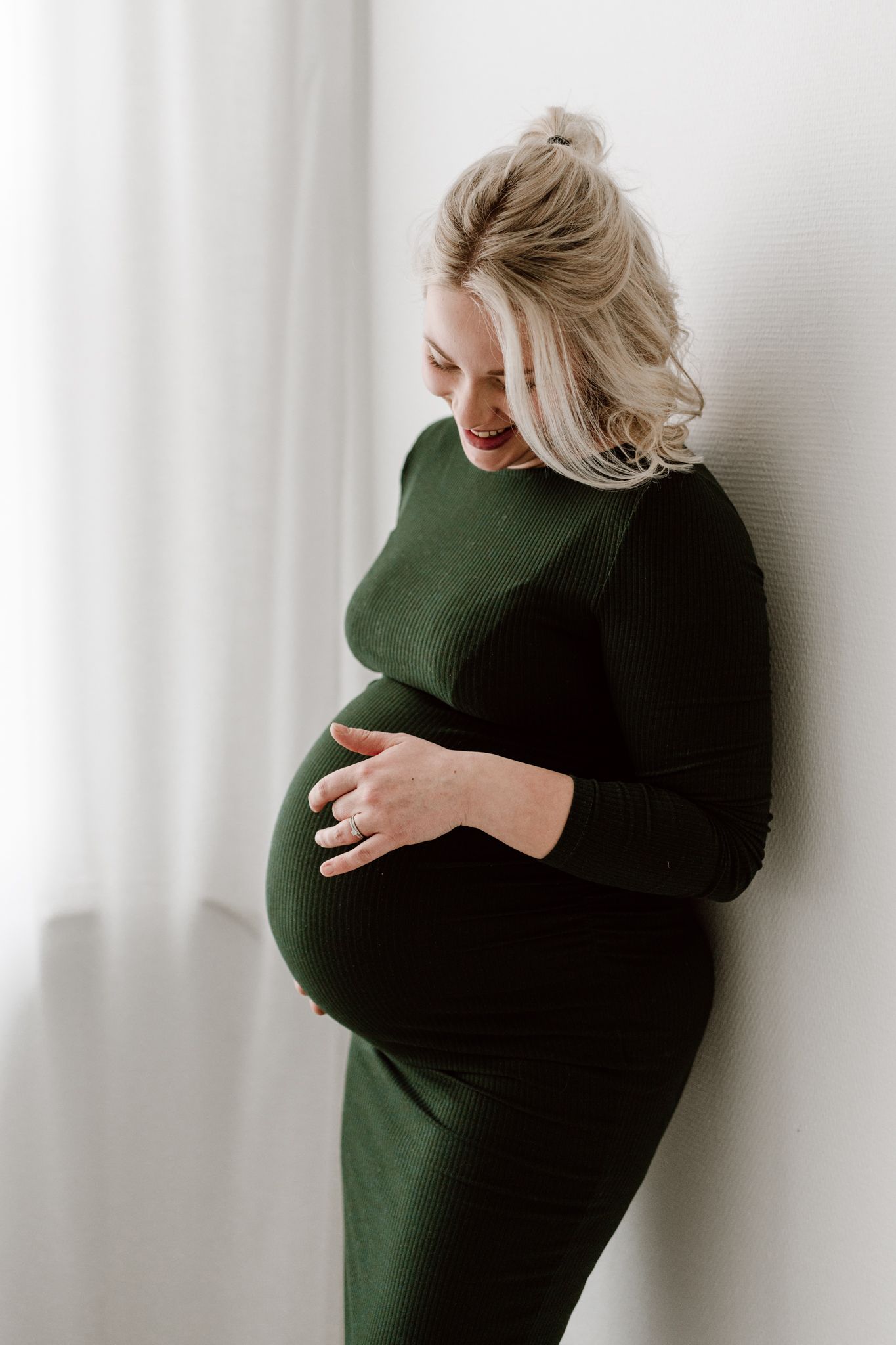 Smiling pregnant woman in dark green ribbed dress cradling her baby bump against white wall.