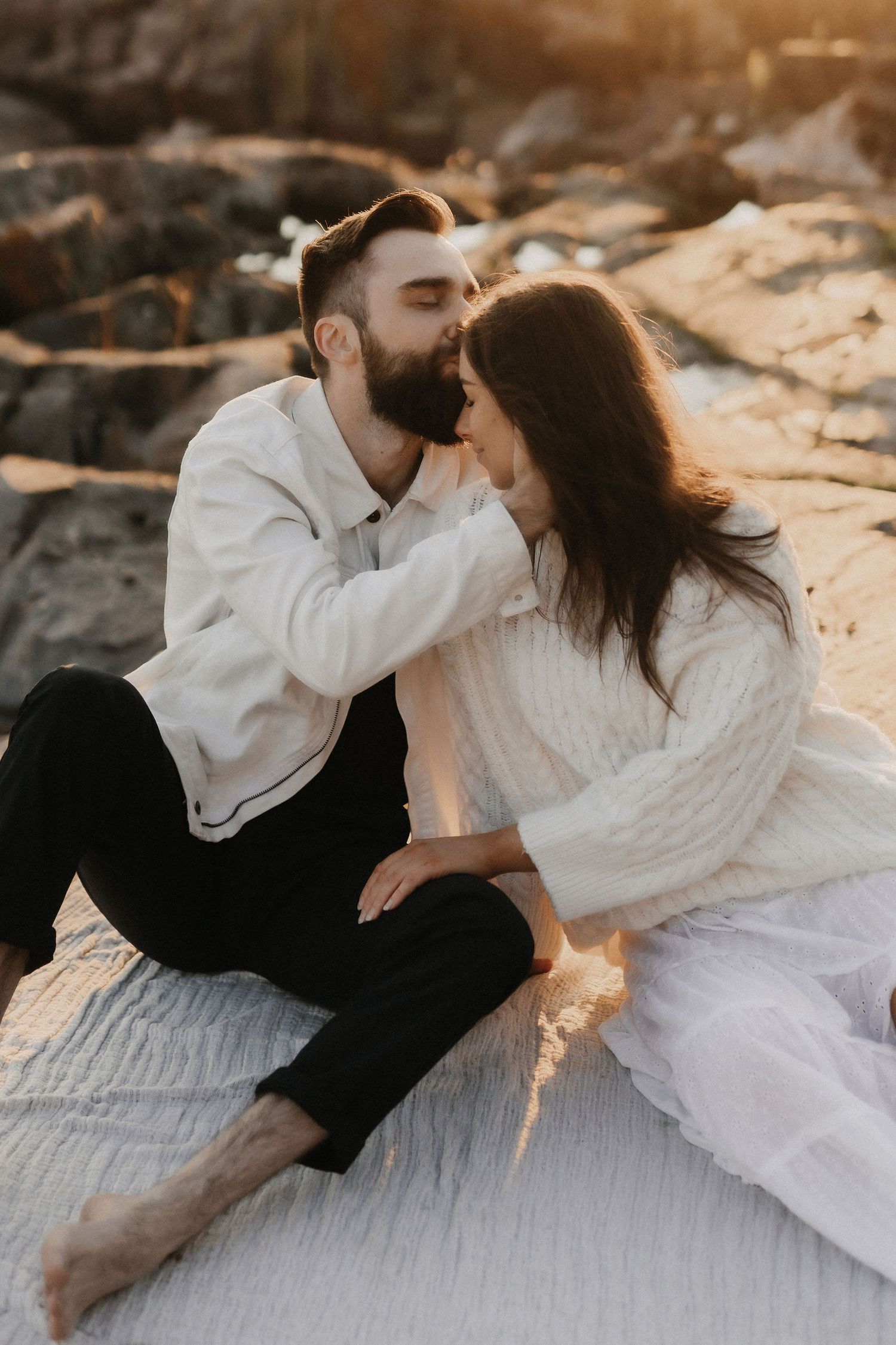 Couple sharing an intimate kiss while sitting on a white blanket on sun-warmed coastal rocks.