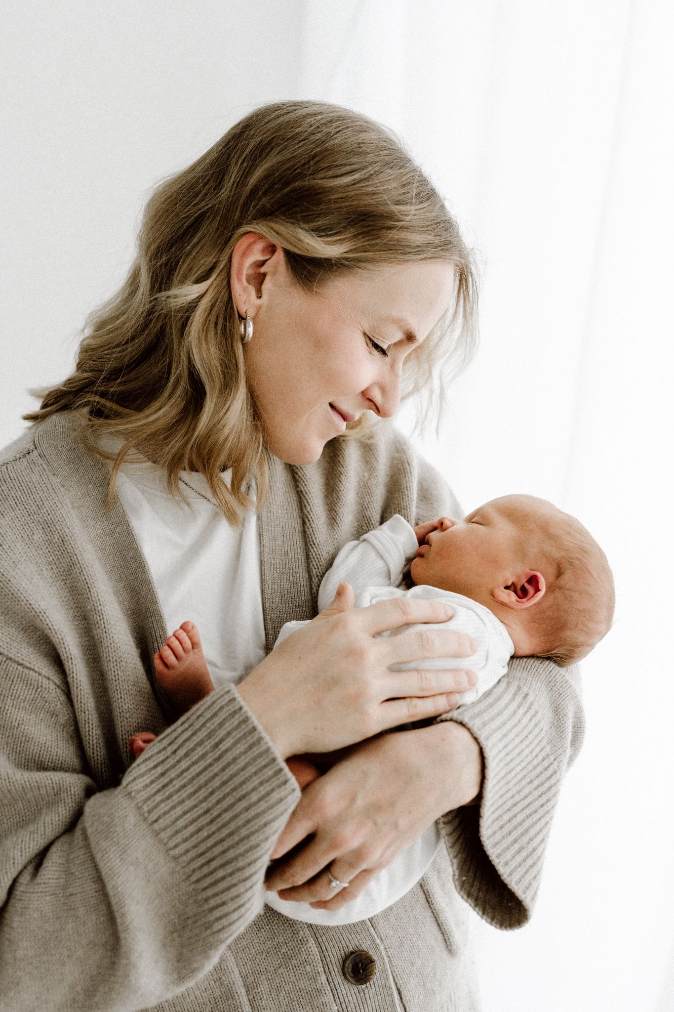 A woman in a neutral cardigan smiles warmly while holding her newborn baby near a bright window.