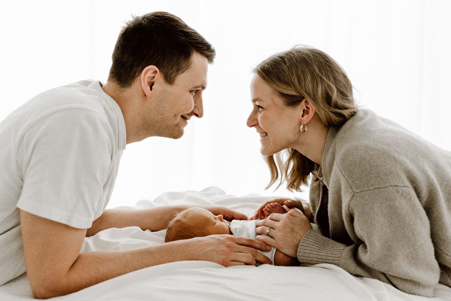 Smiling parents lie on a white bed facing each other, lovingly gazing at their newborn baby between them.
