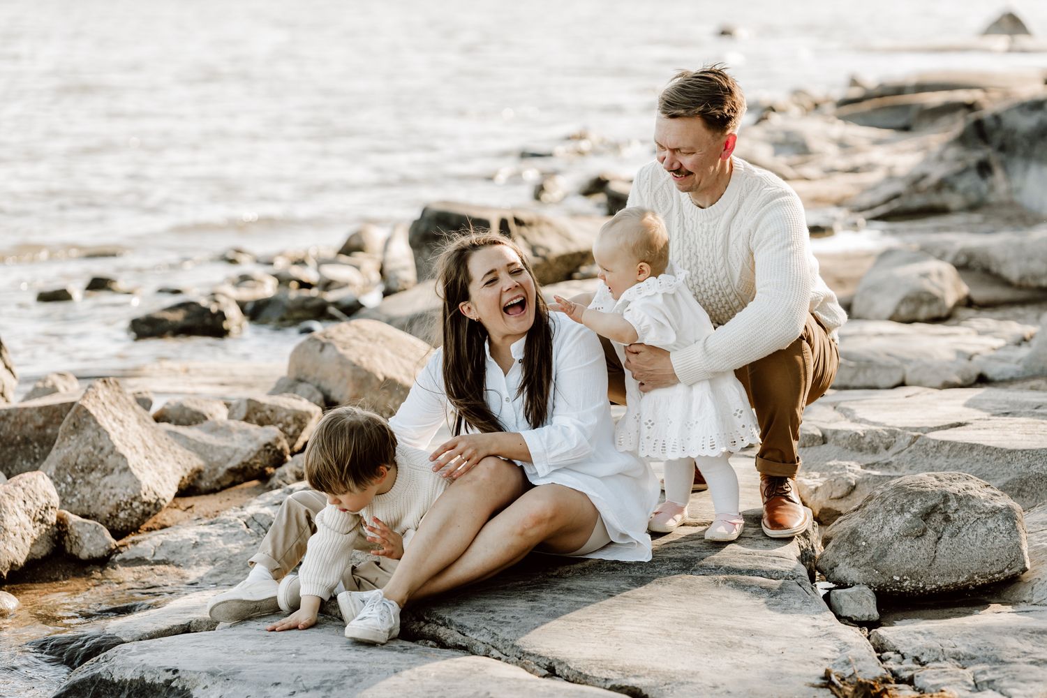A laughing mother sits on rocks by the water as a father and two young children play around her joyfully.