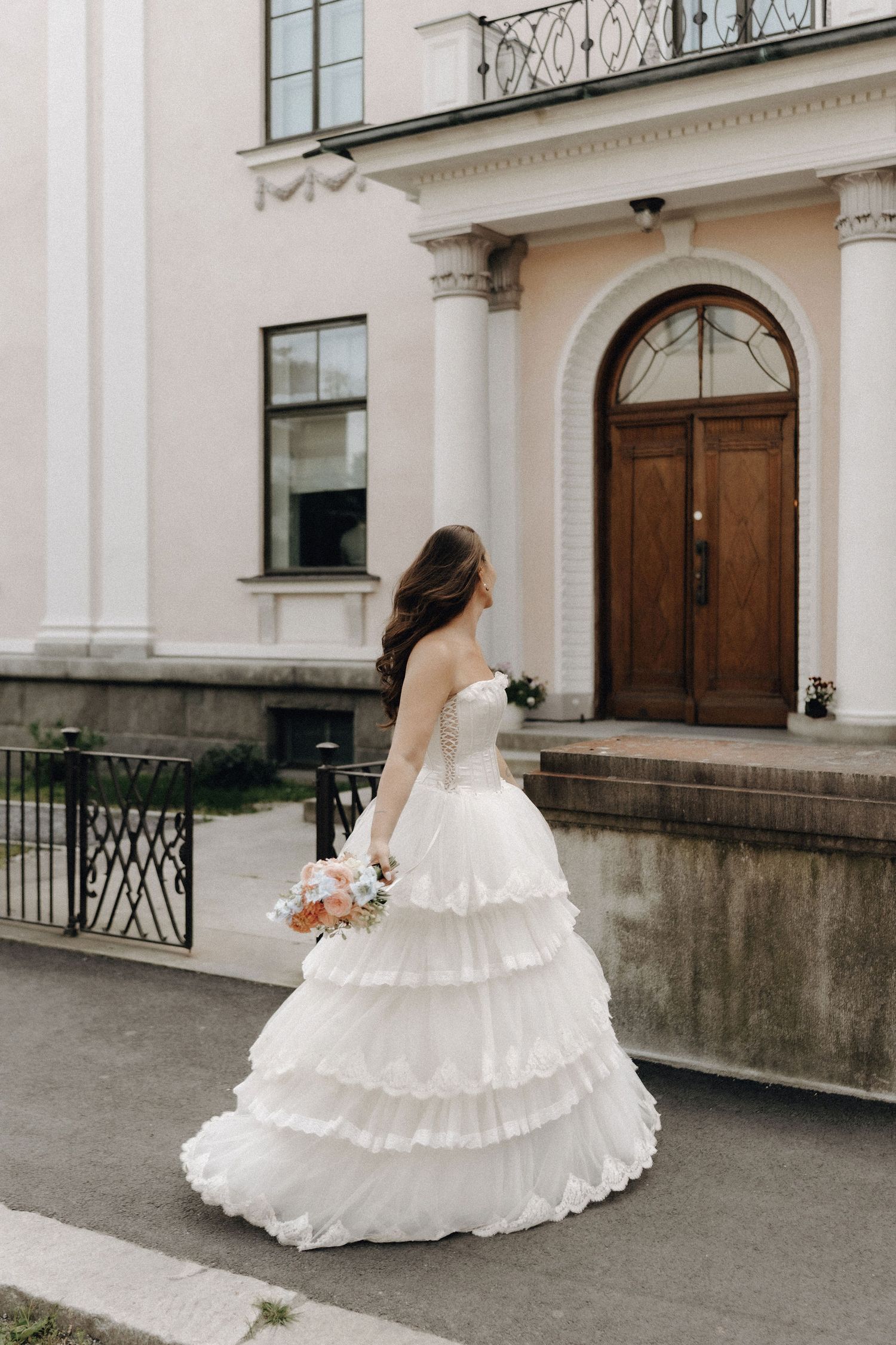 Bride in a strapless white ruffled tiered gown holding a bouquet, standing before an elegant building entrance.