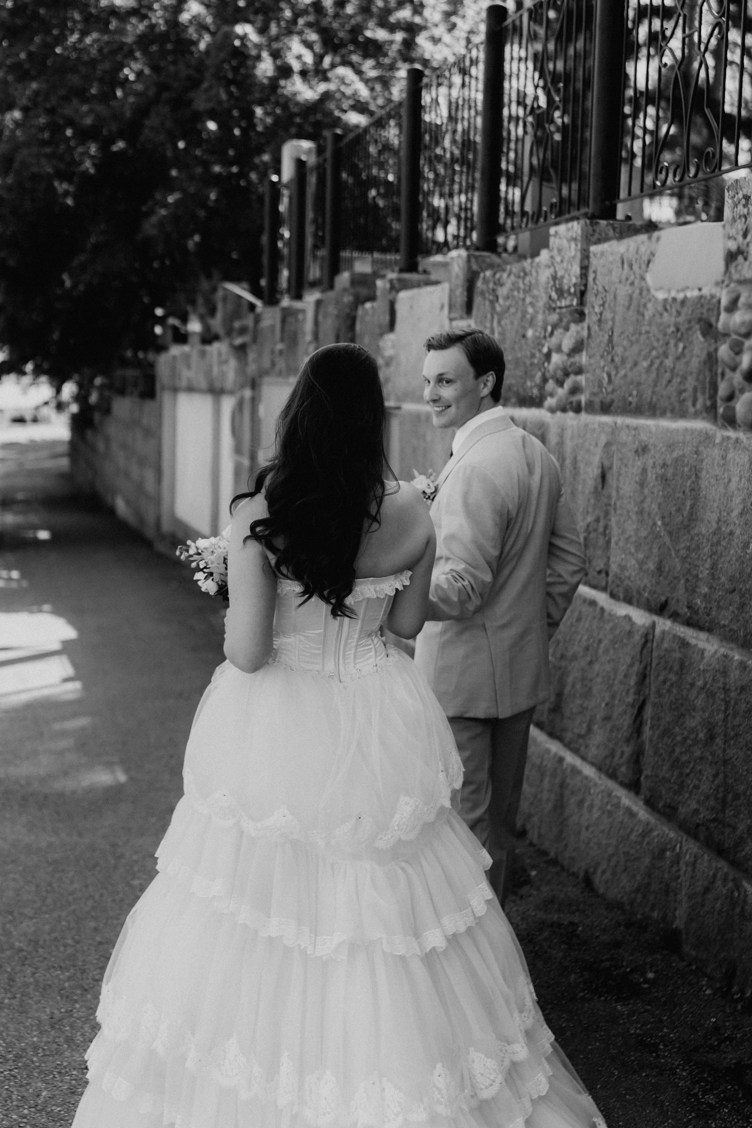 Black and white photo of bride in tiered gown approaching smiling groom along a stone wall pathway.