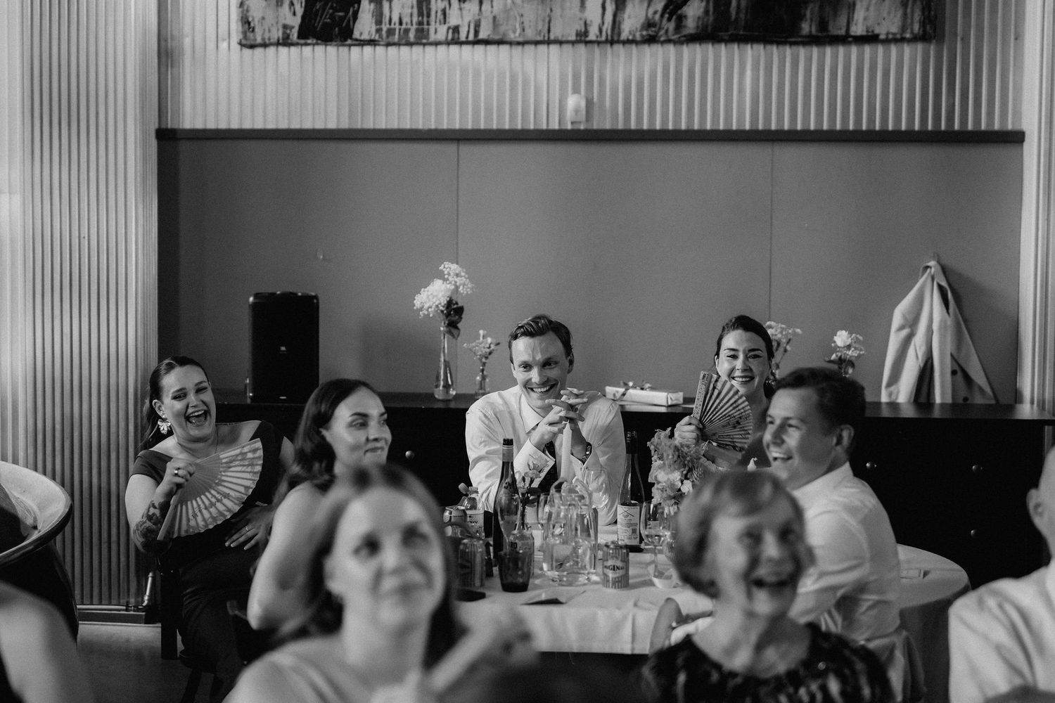Black and white photo of wedding guests laughing during reception speeches at decorated tables.