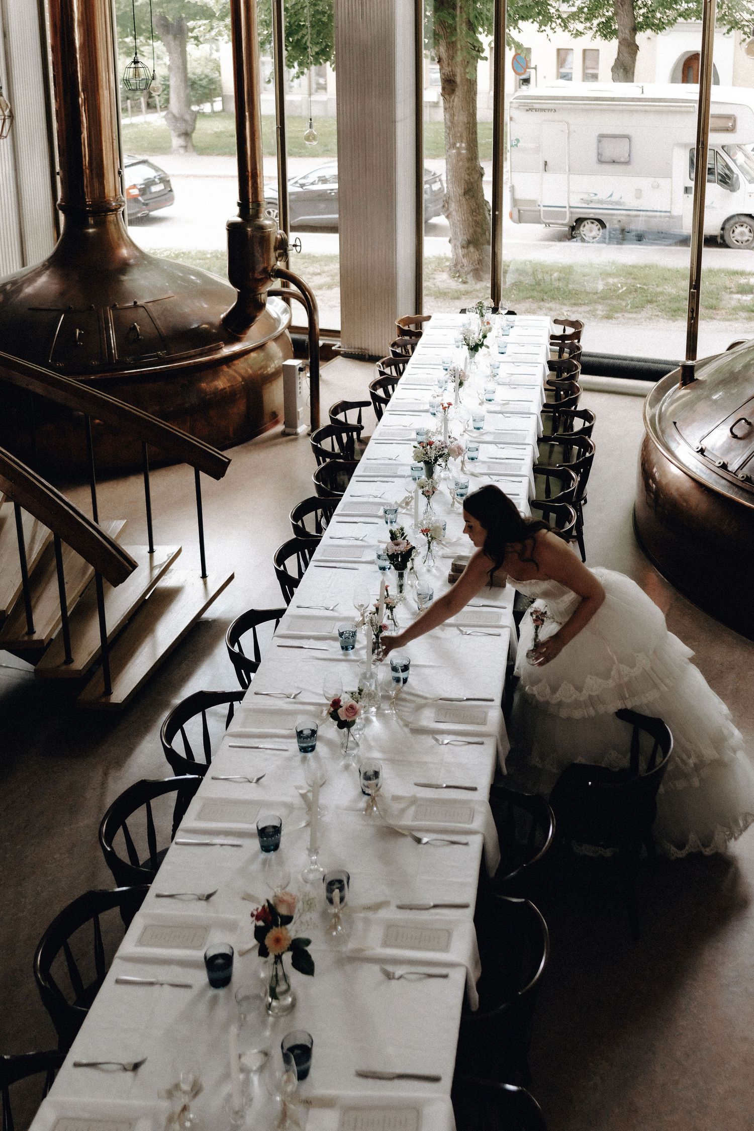 Bride in ruffled white gown arranges long reception table in industrial brewery venue with copper tanks.