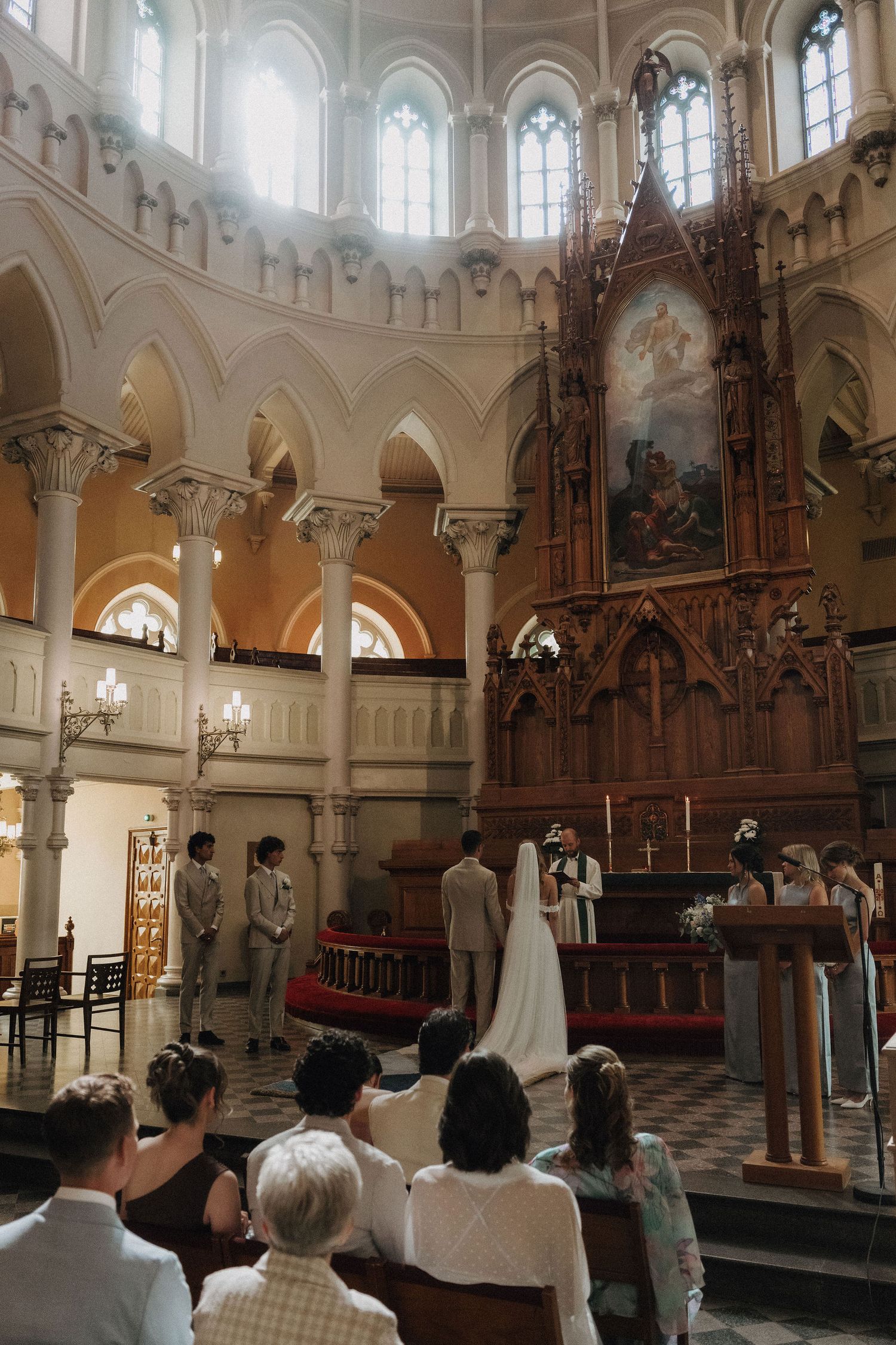 Church wedding ceremony viewed from the side showing bride in white gown and groom at ornate altar with dome ceiling.