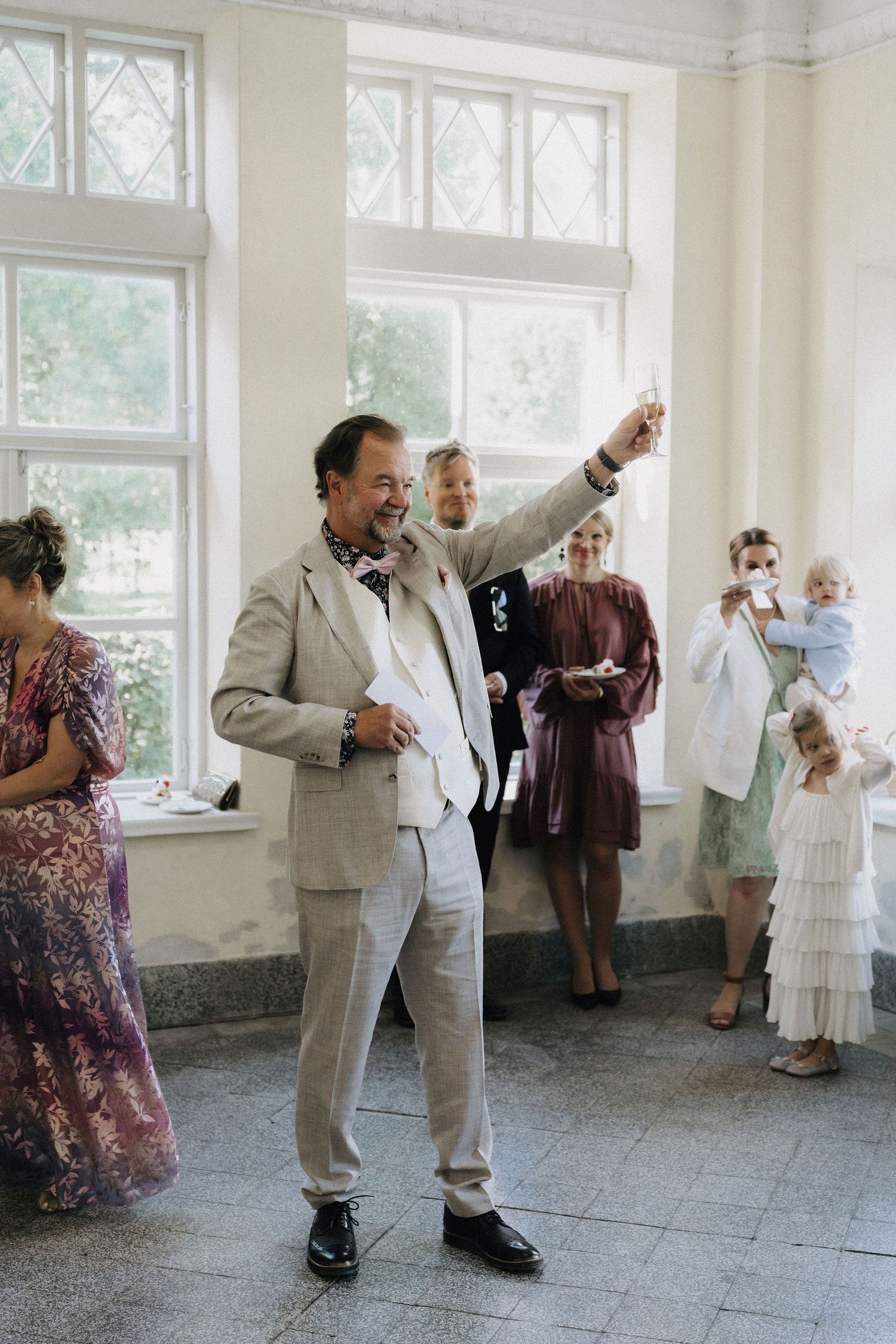 Older man in linen suit raises champagne glass while giving a toast at wedding reception.