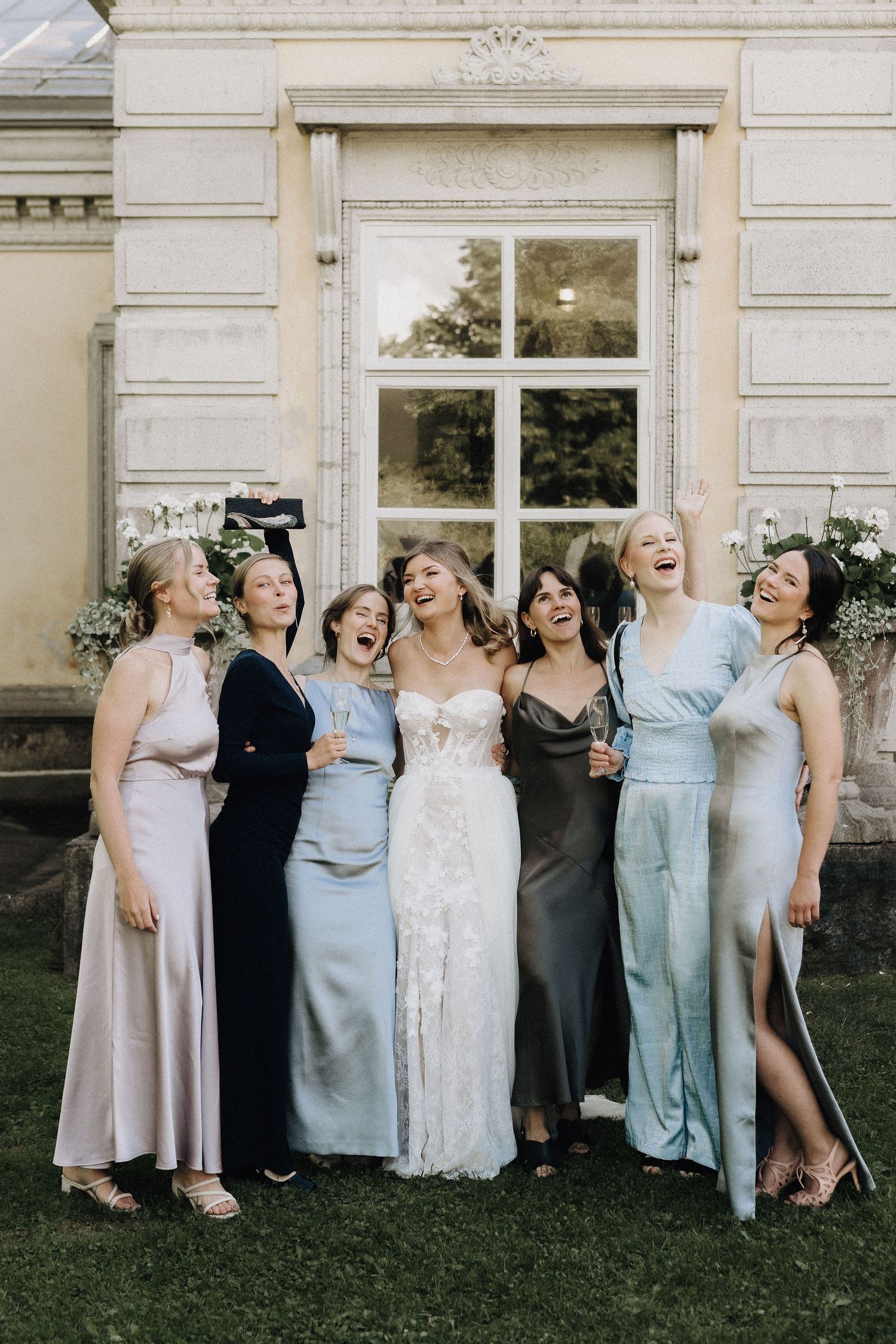 Bride in lace gown laughing joyfully with six bridesmaids in mismatched satin dresses outside a grand building.