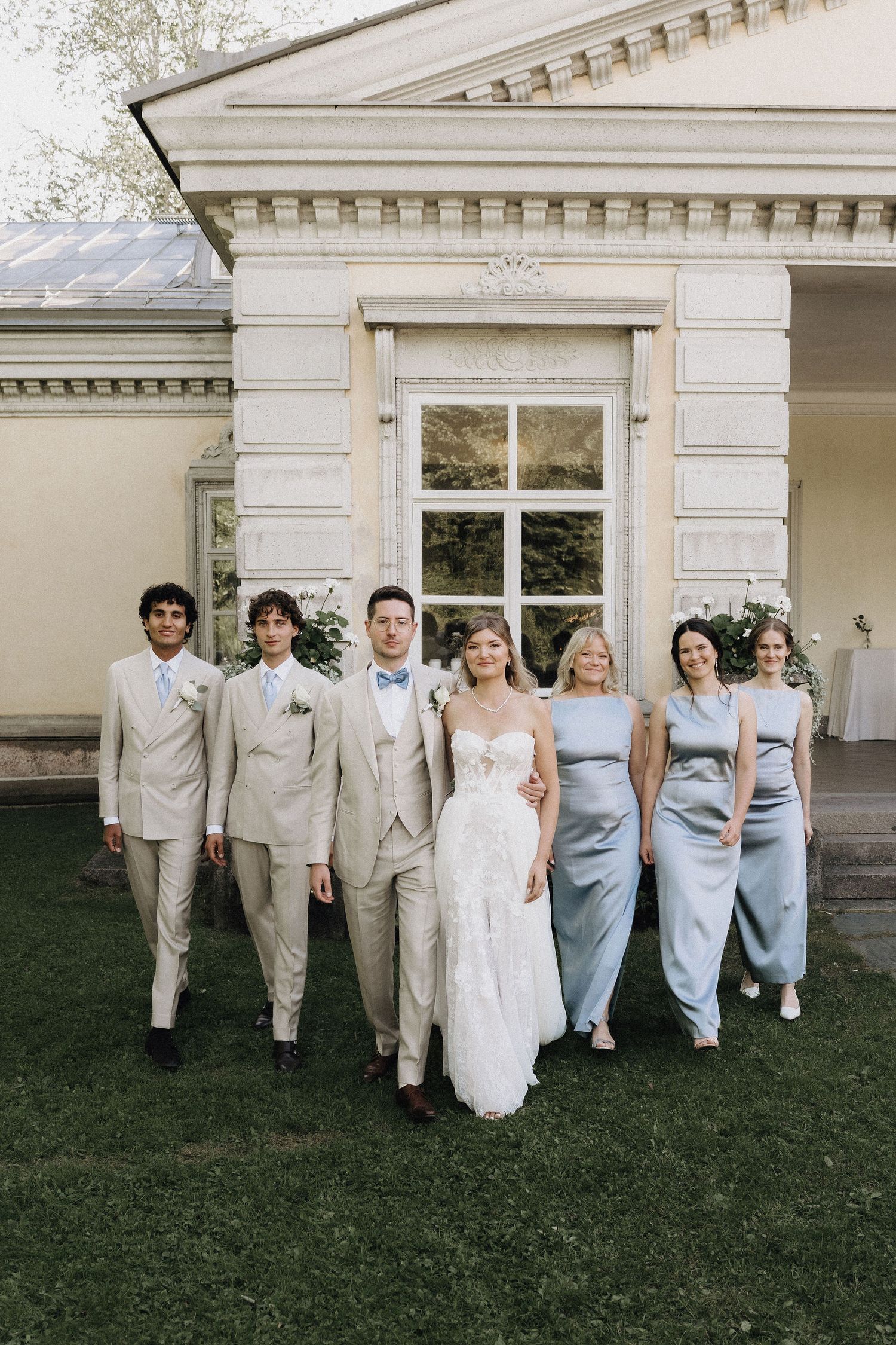 Wedding party poses outdoors in elegant attire, bride in white lace gown with groomsmen and bridesmaids in blue.
