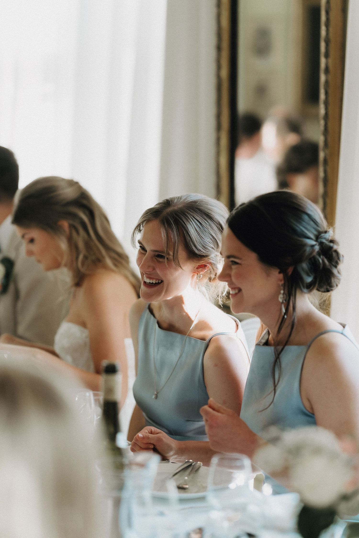 Two smiling bridesmaids in light blue dresses sharing a joyful moment at a wedding reception dinner.