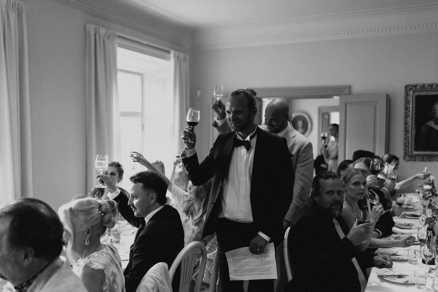 Man in tuxedo raises glass during wedding reception toast while guests celebrate at elegantly set dining table.