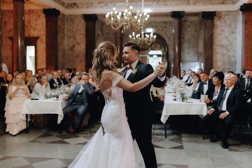 Bride in lace mermaid gown and groom dancing elegantly at wedding reception in marble hall.
