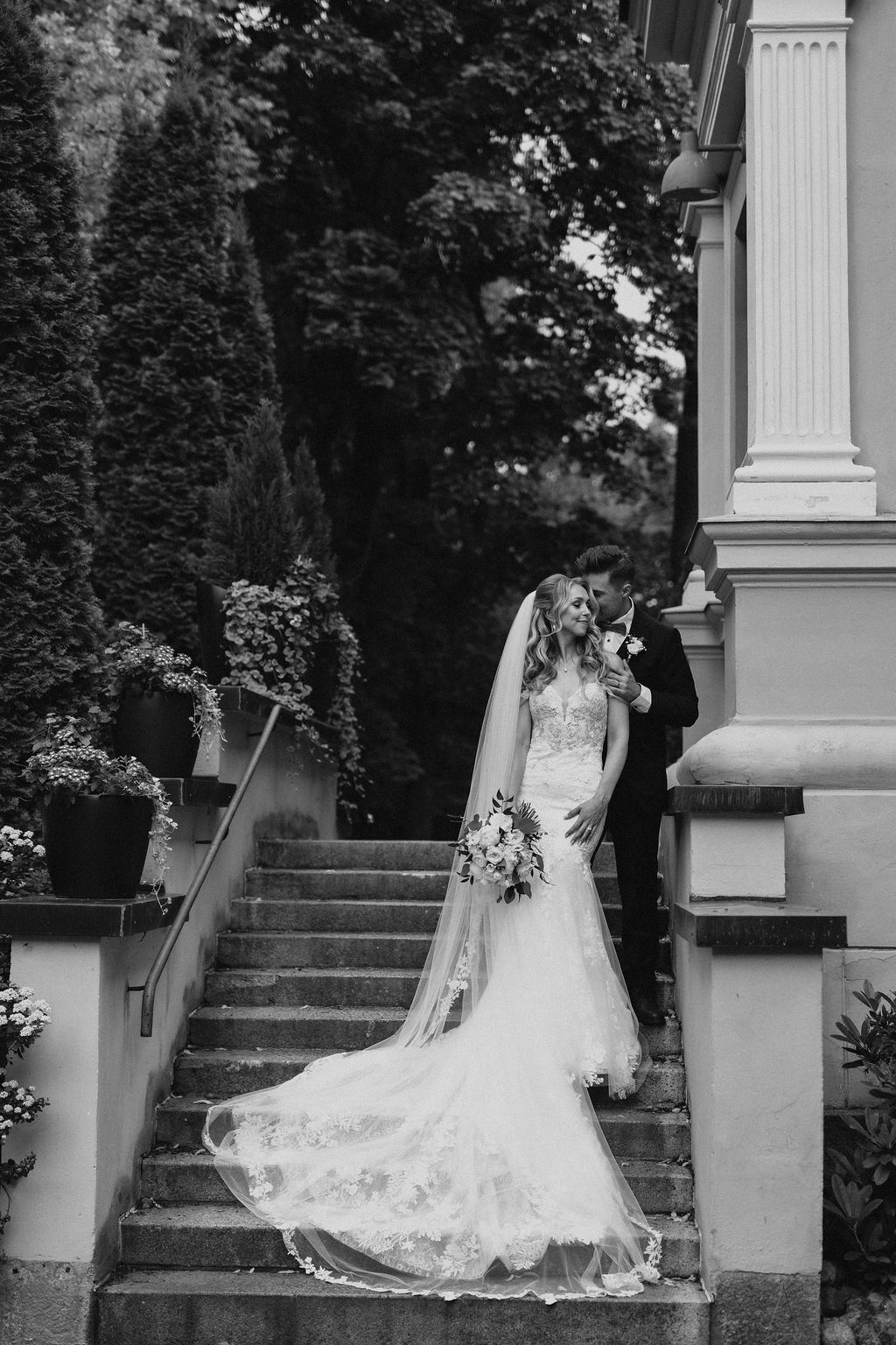 Black and white photo of bride and groom sharing a kiss on grand stone staircase outdoors.