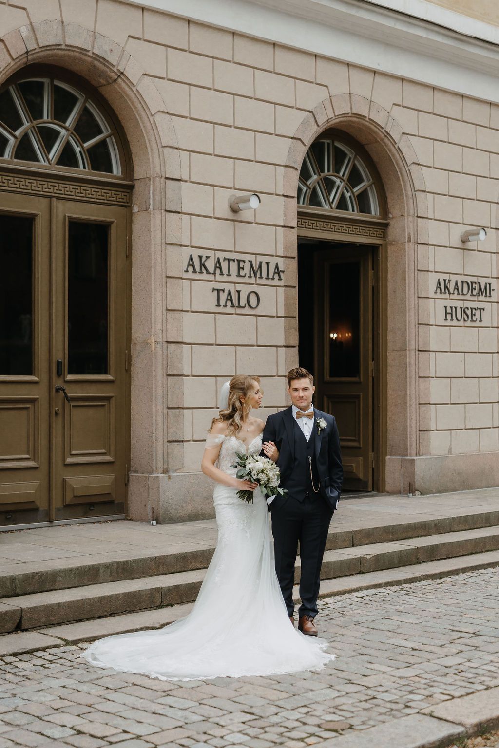 Bride and groom posing in front of Akatemia-Talo building on cobblestone street, bride holding white bouquet.