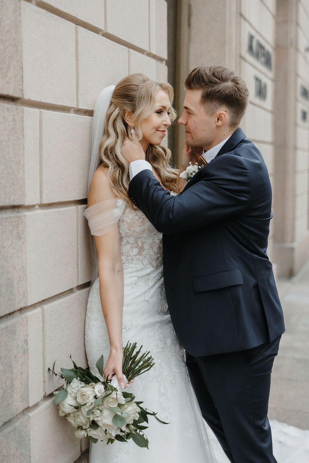 Groom whispering to smiling bride leaning against stone wall, bride holding elegant white rose bouquet.