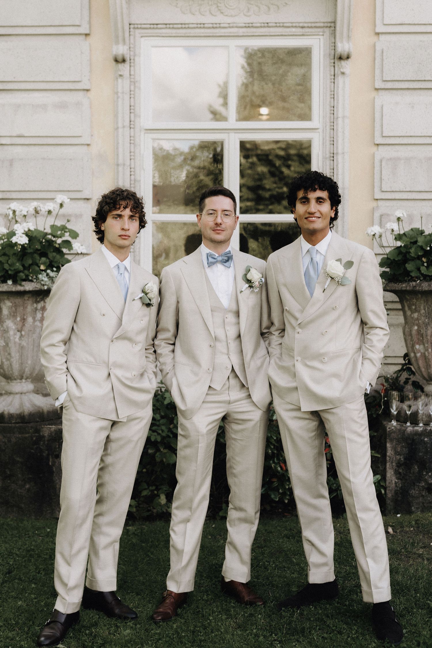 Three groomsmen in matching cream suits with boutonnieres standing before elegant building facade.