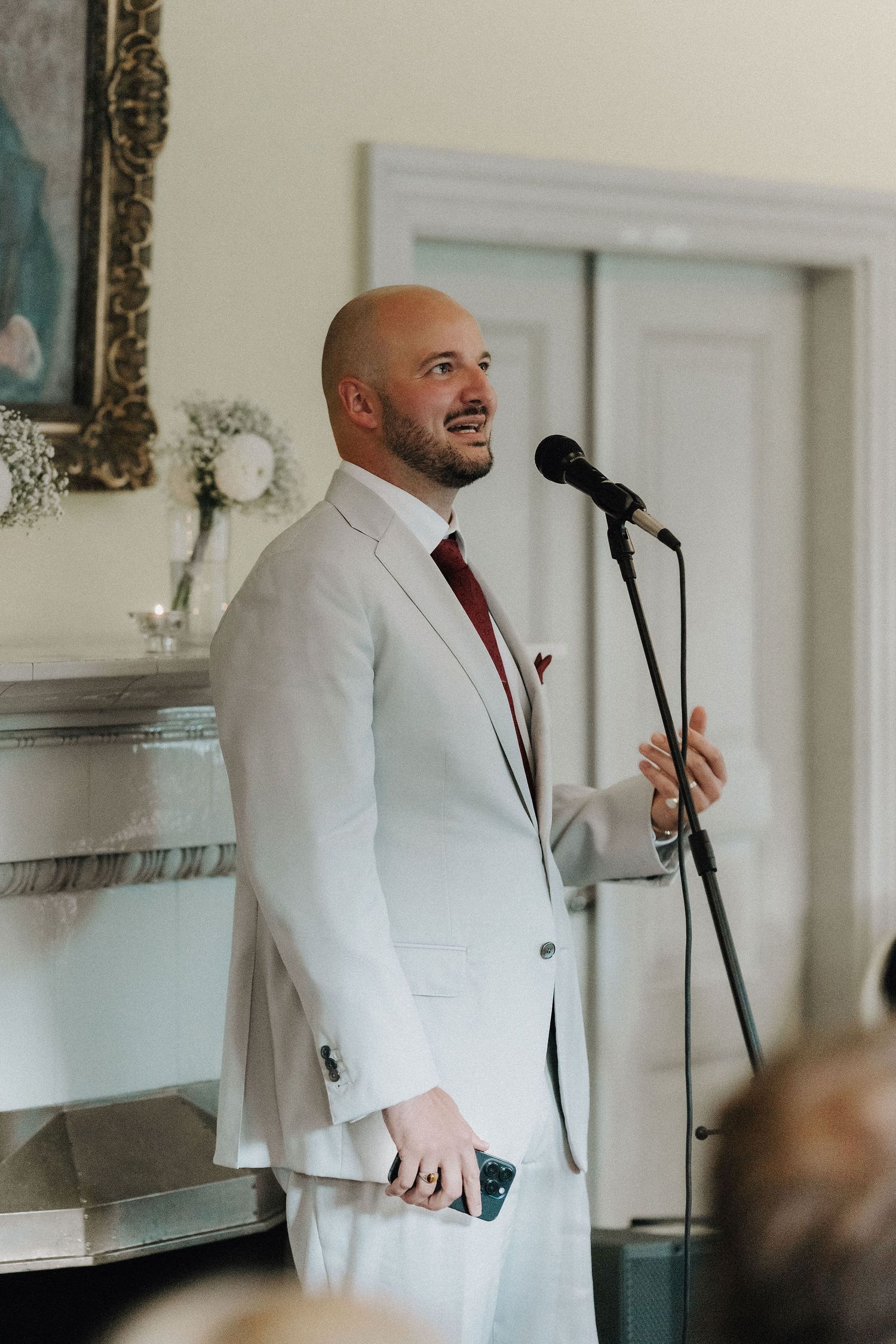 Bald man in white suit with red tie giving speech at microphone during wedding reception.