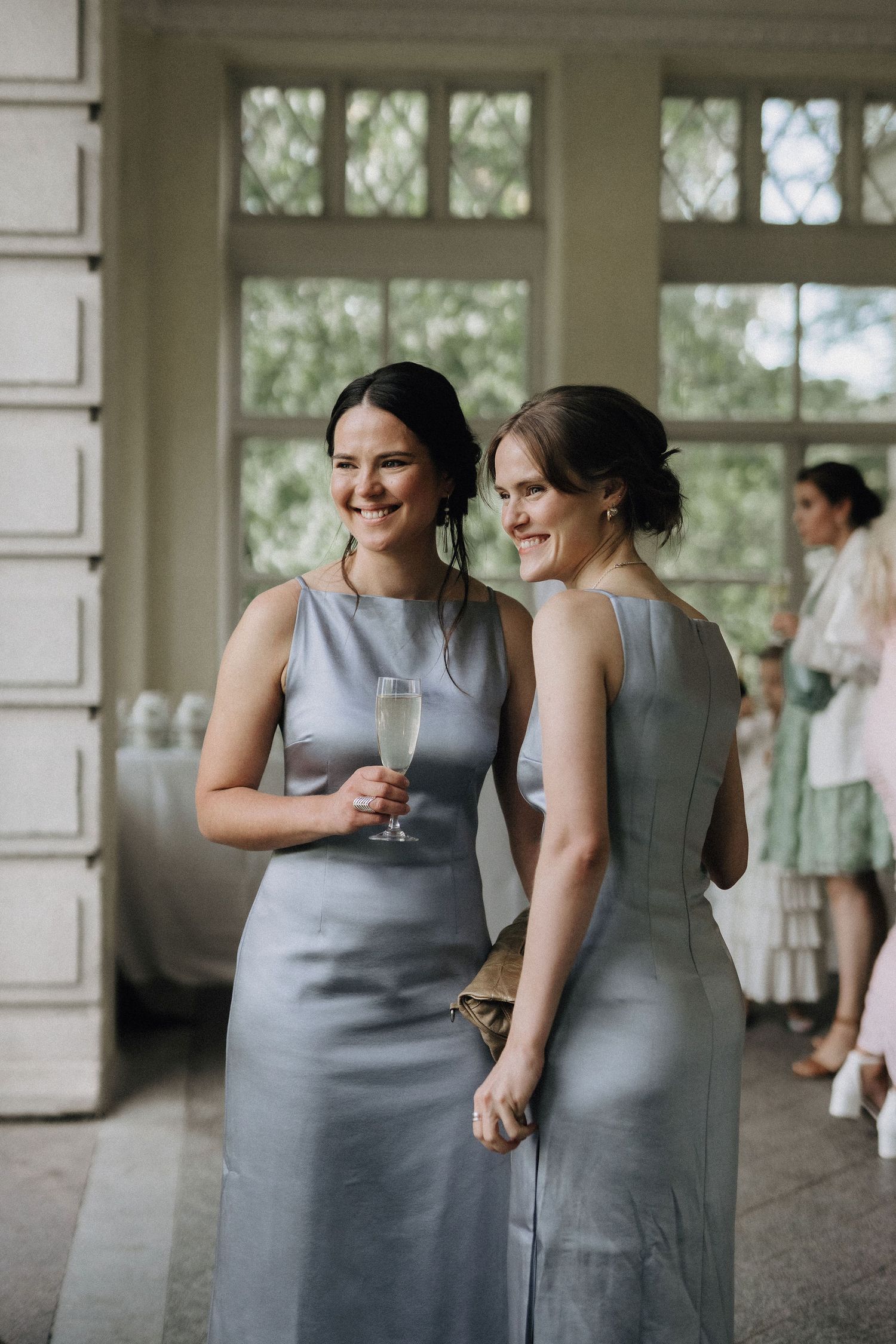 Two bridesmaids in silver satin dresses smiling and holding champagne glasses at a wedding reception.