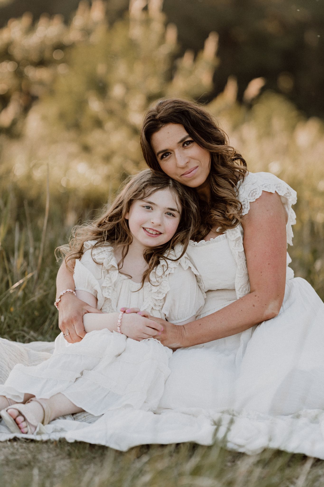 A mother and daughter in white dresses sit together smiling on a blanket in a golden field.