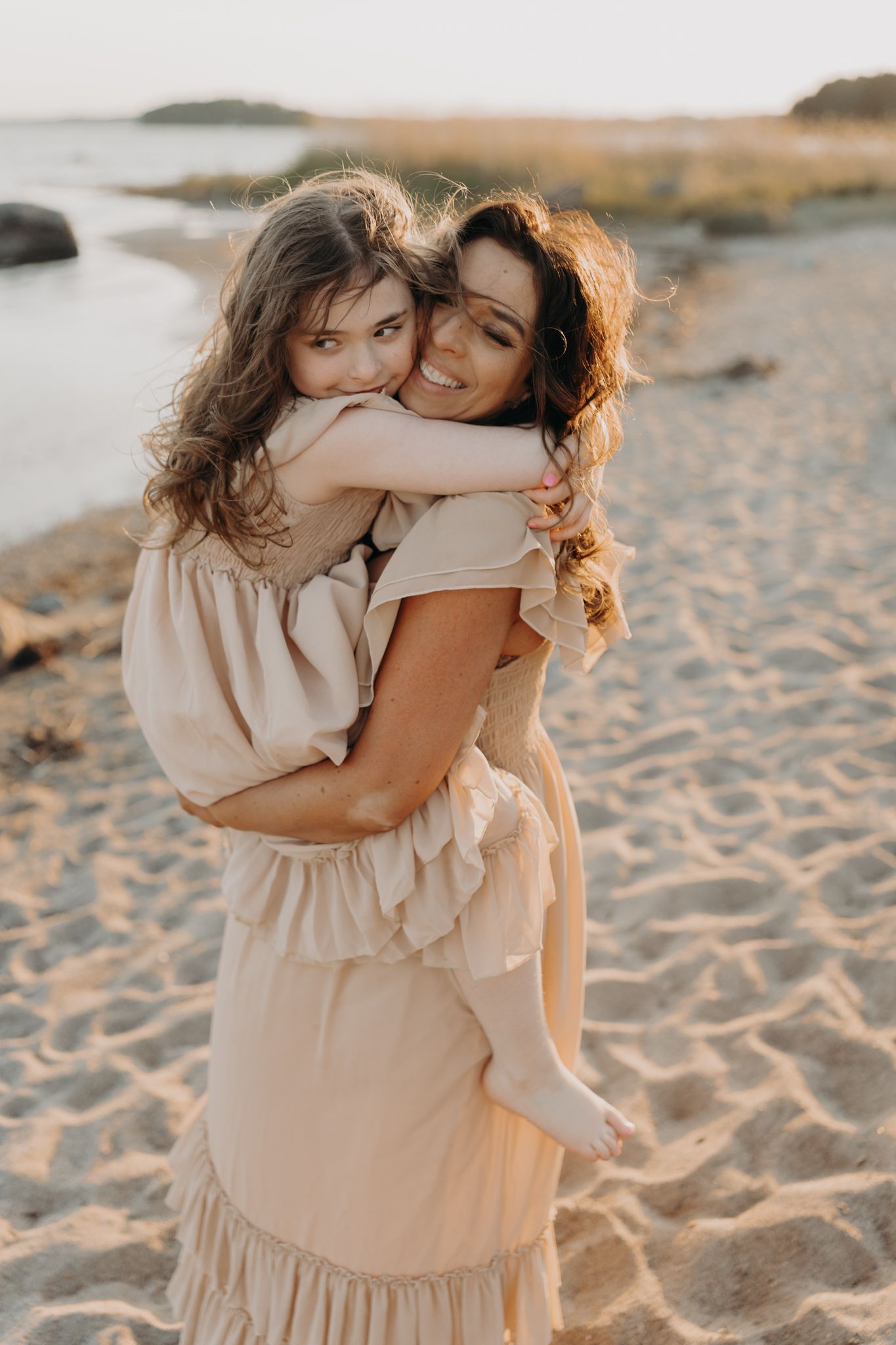 Mother in beige ruffled dress hugging young daughter on sandy beach at sunset.