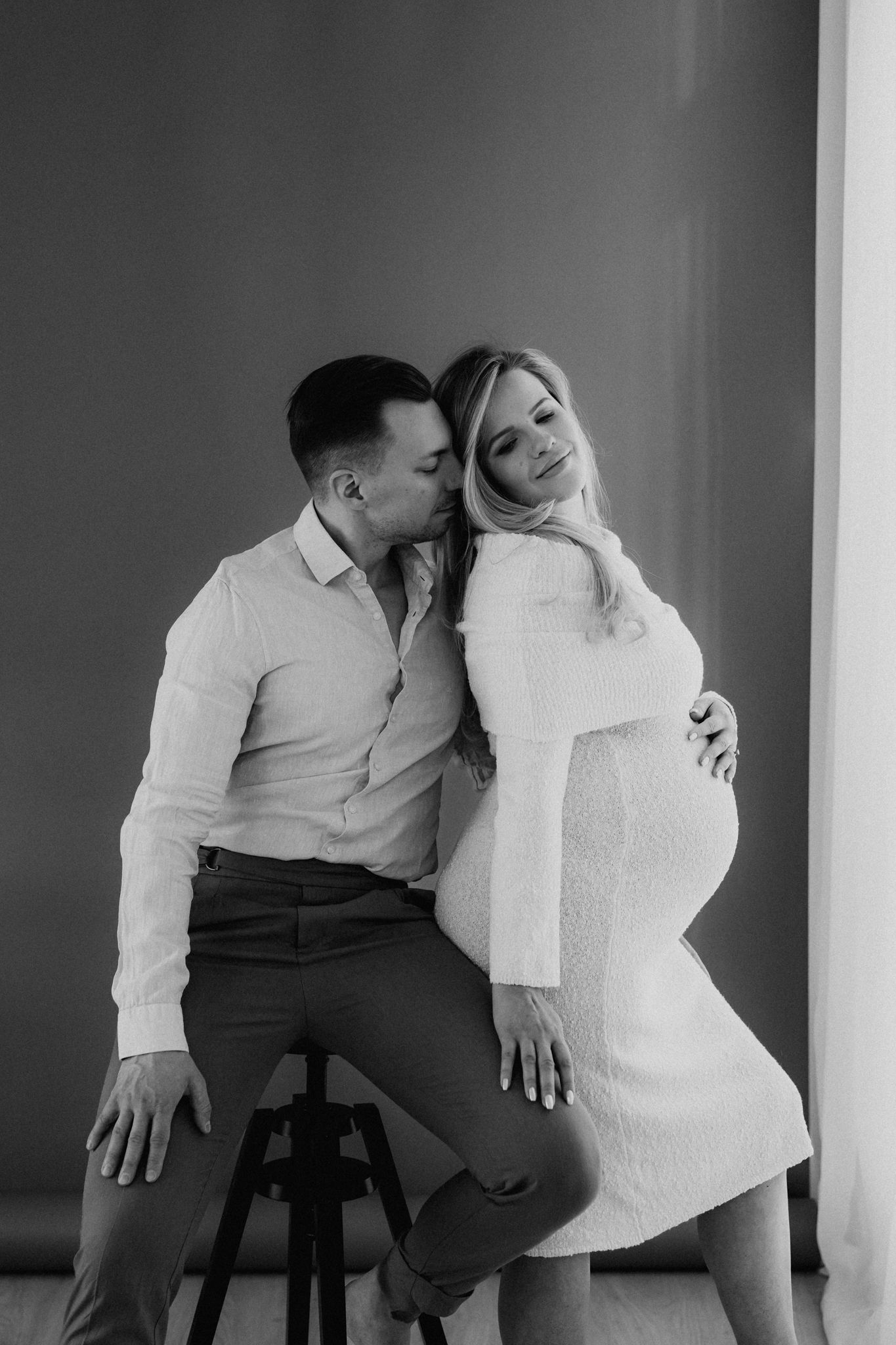 Black and white photo of couple, man kissing pregnant woman in white dress seated on stool.