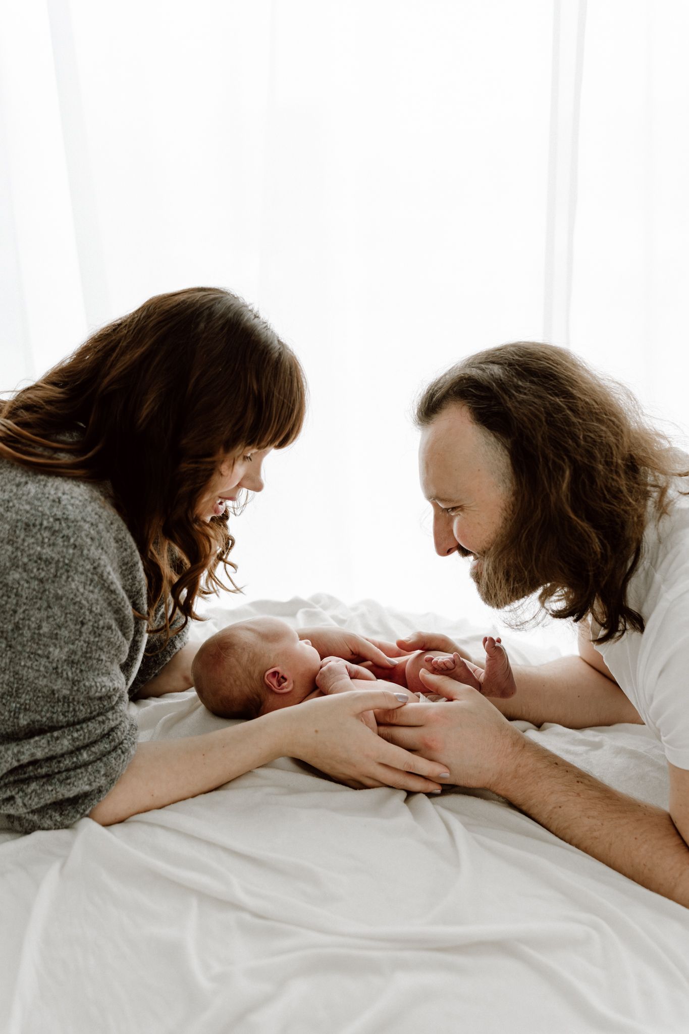Parents lying on white bedding, smiling lovingly at their newborn baby between them.