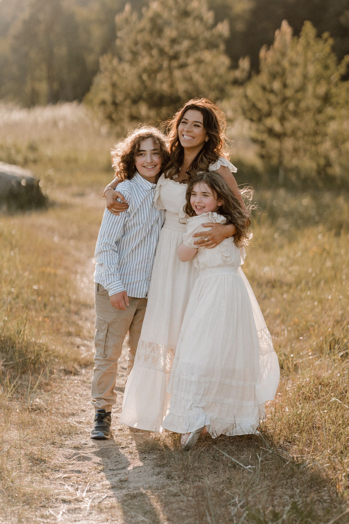 A happy mother in a white dress embraces her two children on a sunlit nature path.