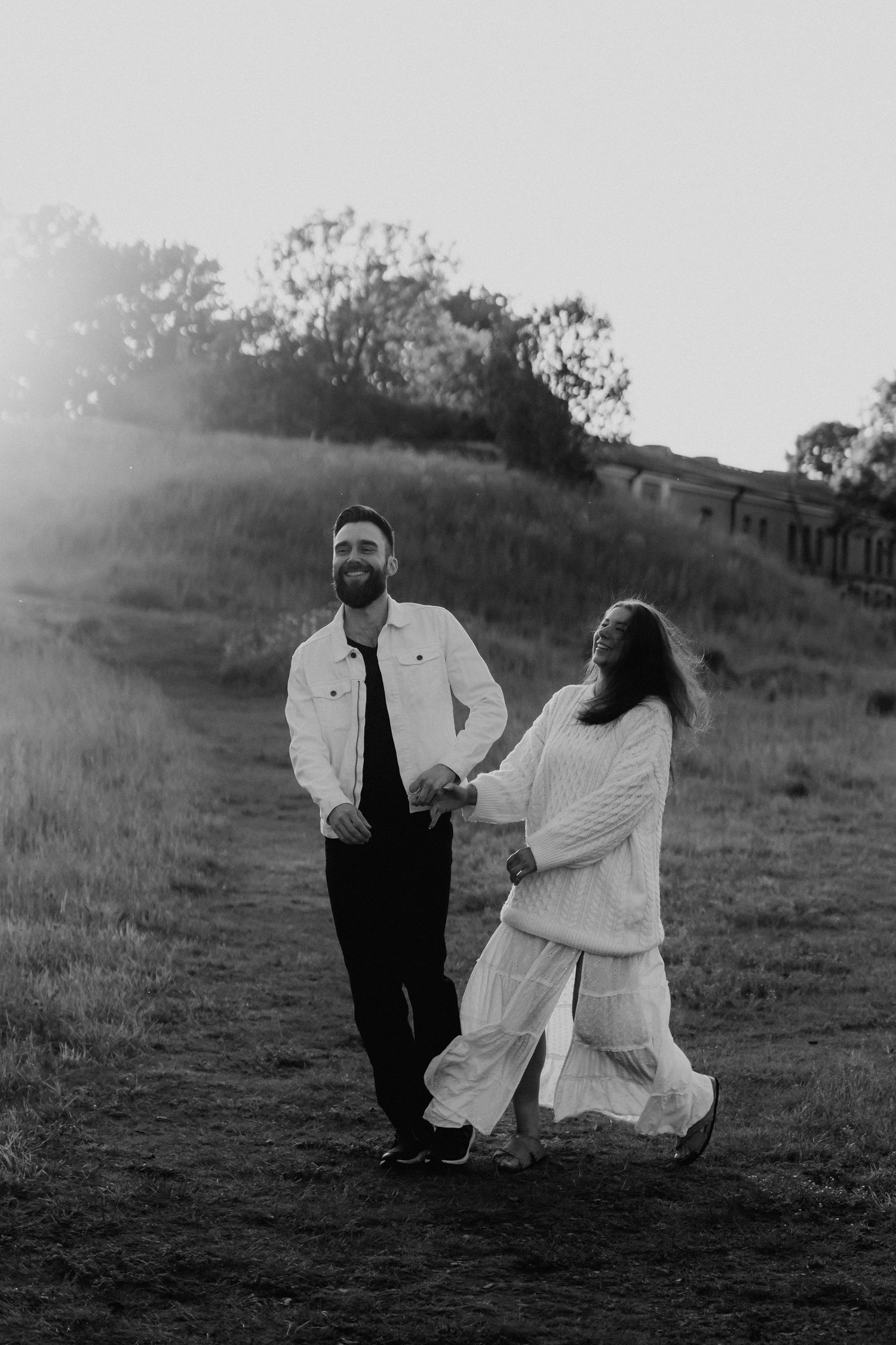Black and white photo of a laughing couple holding hands on a grassy hillside path at dusk.